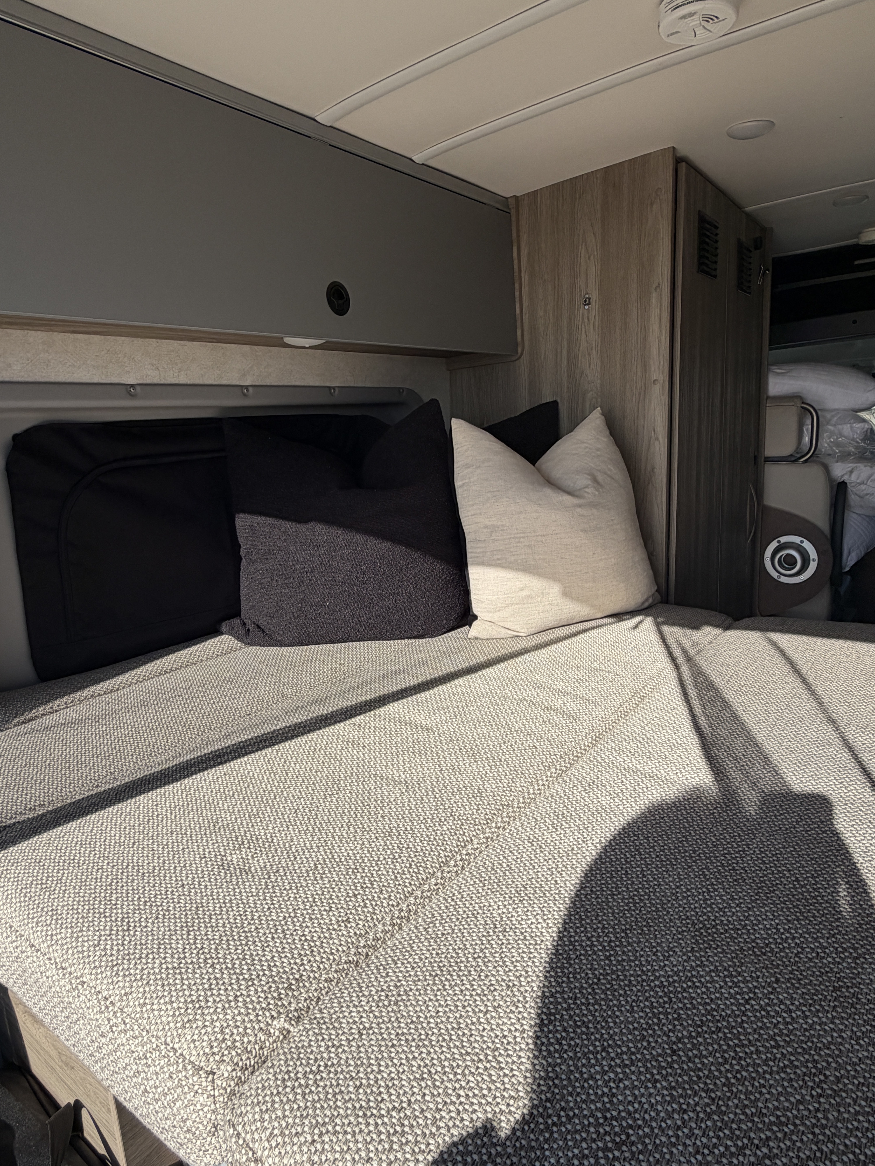 RV rental interior: A sunlit bed with black and cream pillows, gray and wood-toned overhead storage, and a smoke detector.. Winnebago Solis 2025