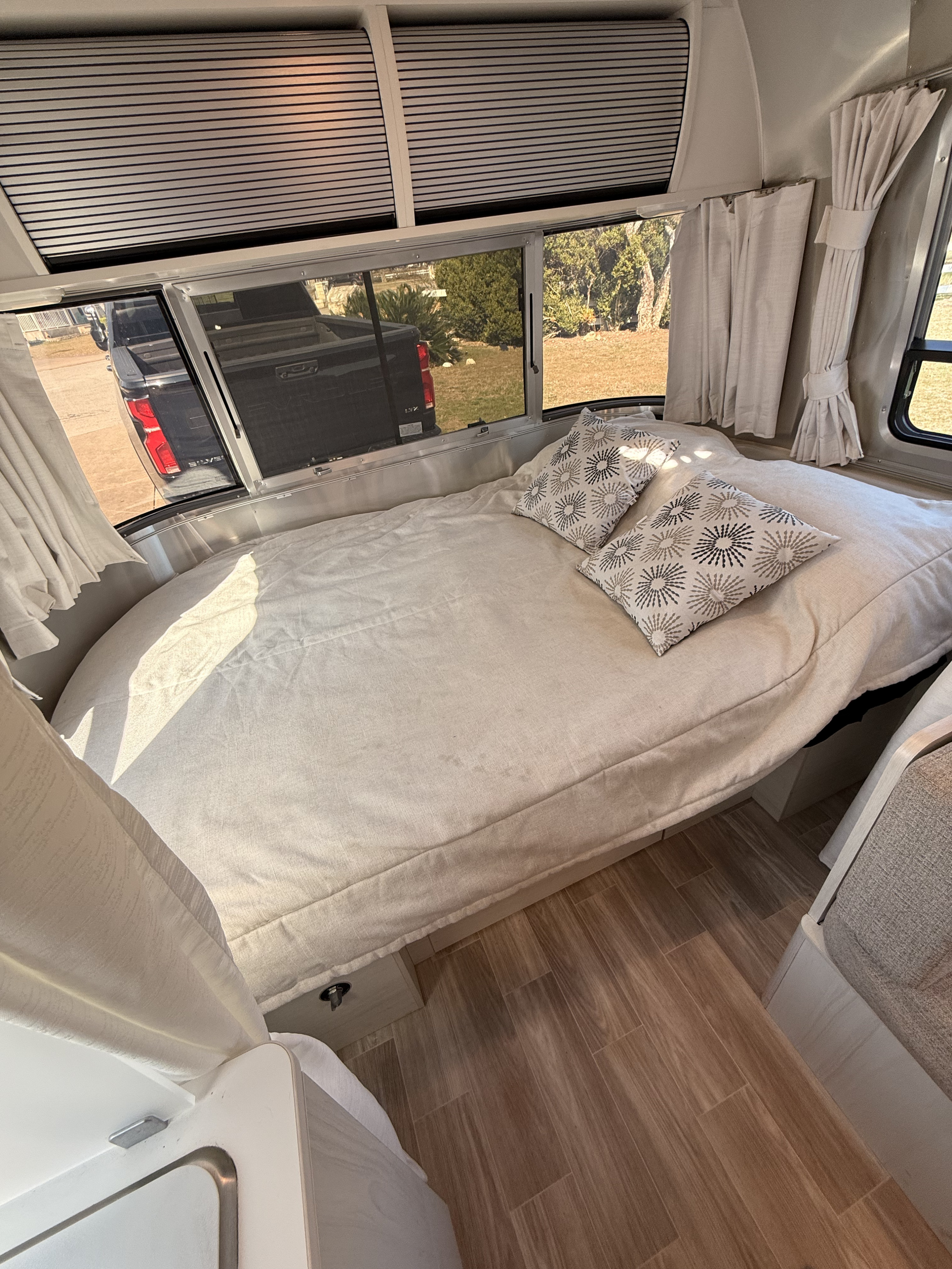 Cozy RV rental bedroom with a comfortable bed, patterned pillows, windows displaying outdoor scenery, and light wood floors.. Airstream Bambi 2024