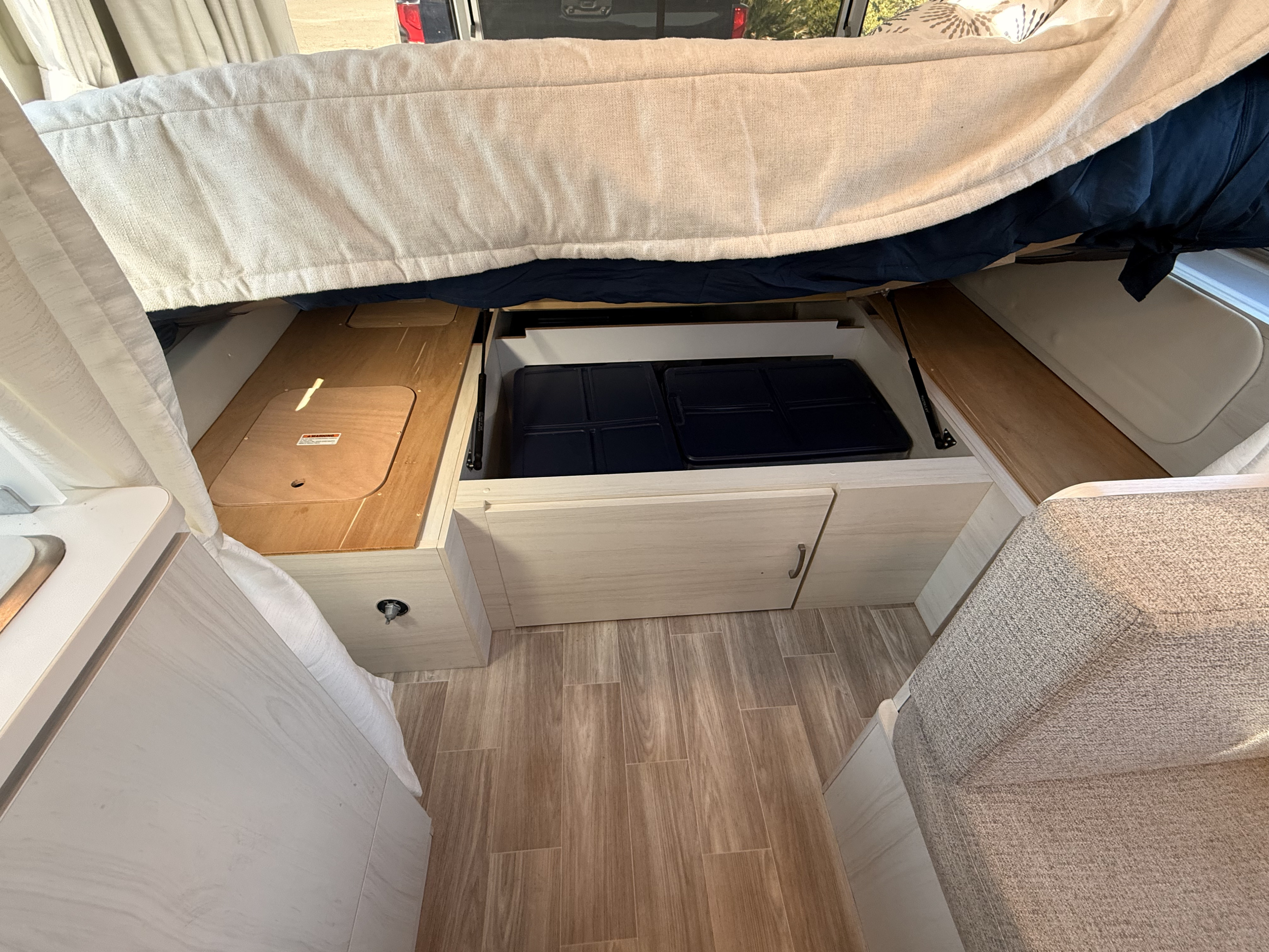RV rental: Interior shows lifted bed revealing under-storage with cooking griddles, light cabinets, wood-look floor, and seating.. Airstream Bambi 2024