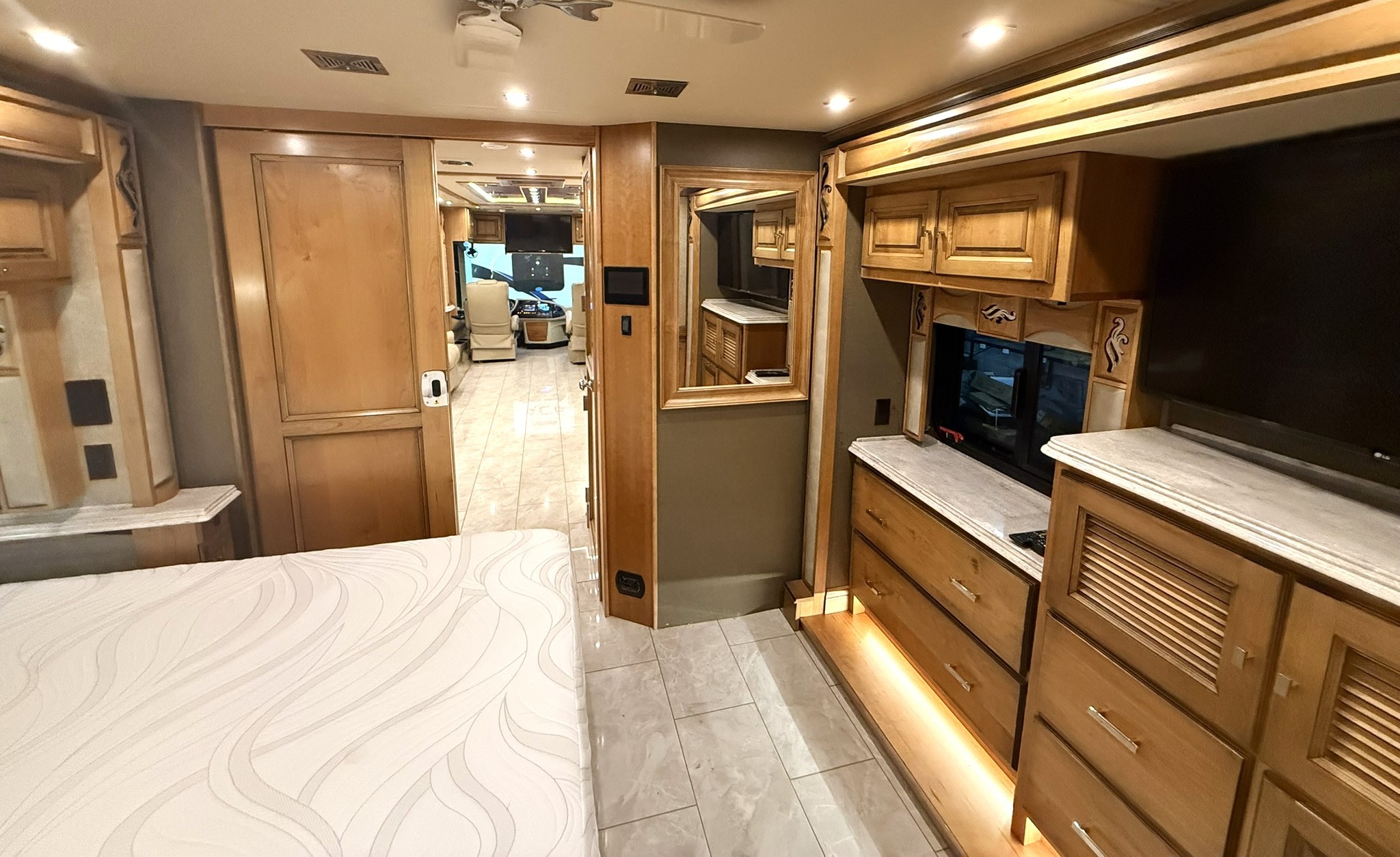 Spacious rv rental bedroom with a comfy mattress, wood cabinetry, TV, and mirror, leading to the living area. Tiled floor.. Tiffin Phaeton 2020