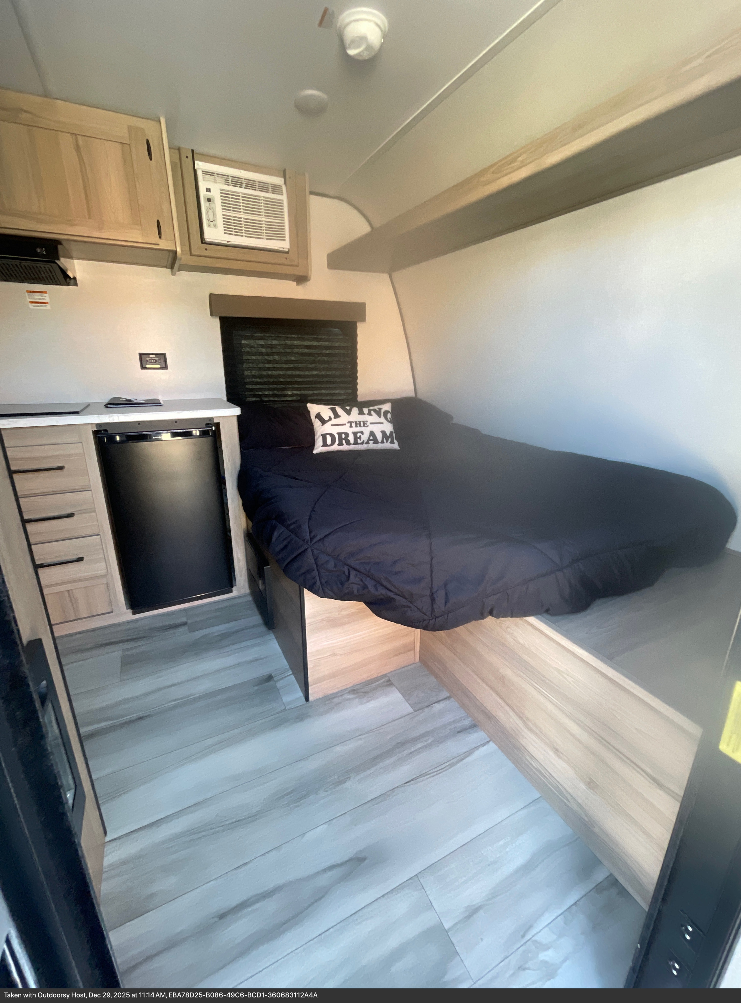 This RV rental interior features a cozy bed with a "LIVING THE DREAM" pillow, mini-fridge, AC, and storage.. Keystone Coleman 13B 2026