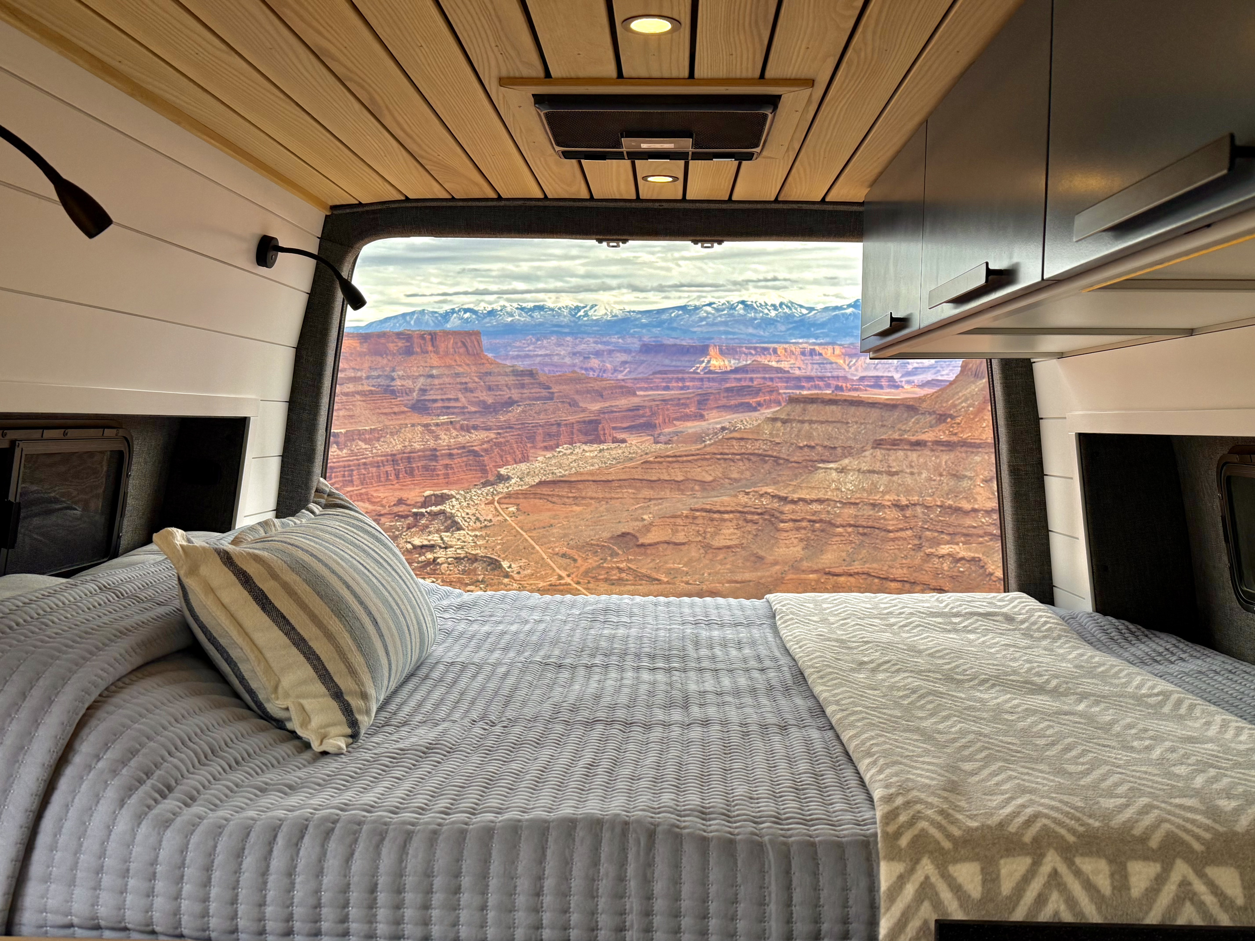 Inside a cozy RV rental, a neatly made bed offers panoramic views of red rock canyons & distant snow-capped mountains.. Mercedes-Benz Sprinter 2023