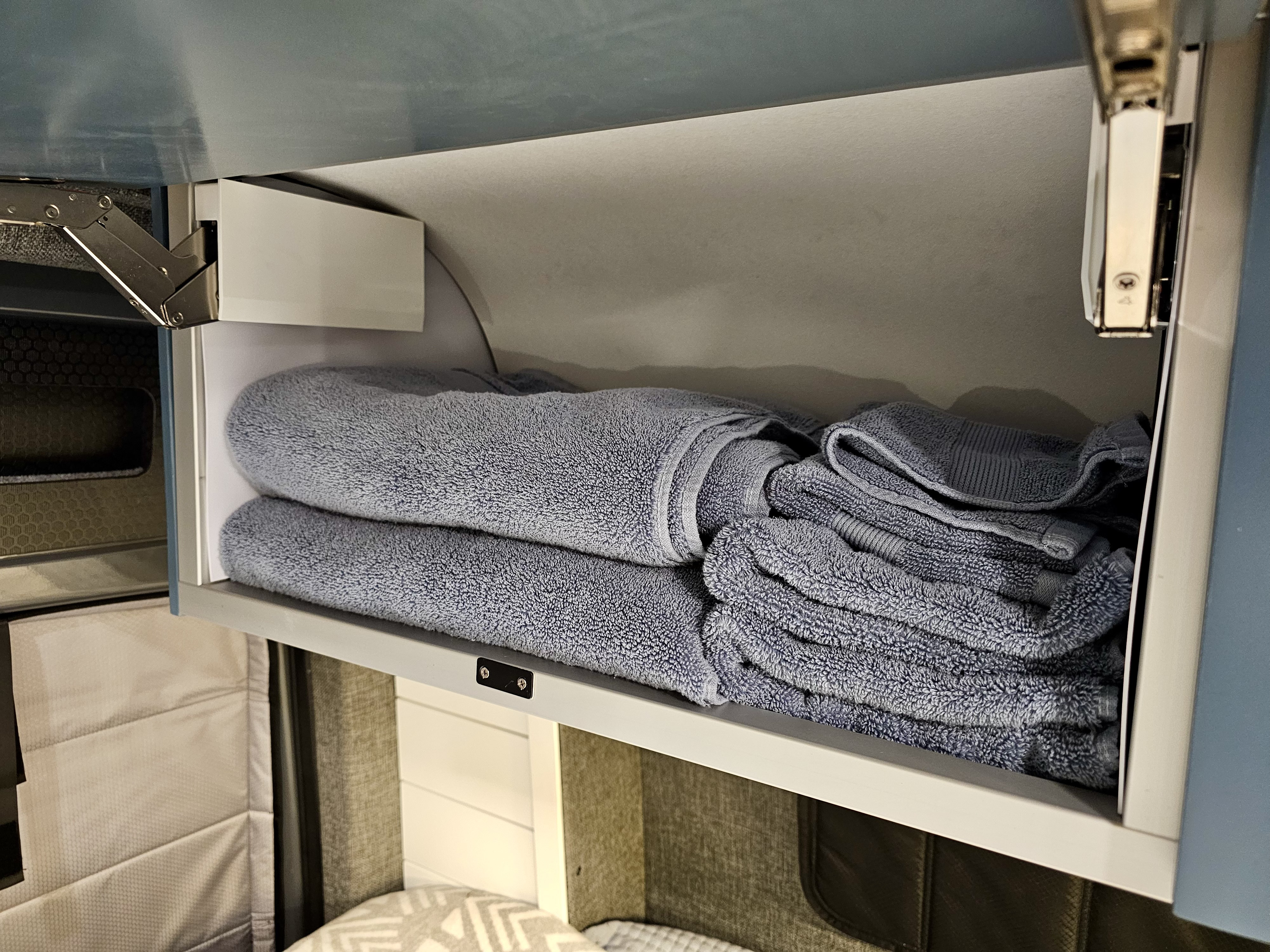 Soft blue towels organized in an overhead cabinet in an RV rental. Practical, space-saving storage for travel essentials.. Mercedes-Benz Sprinter 2023