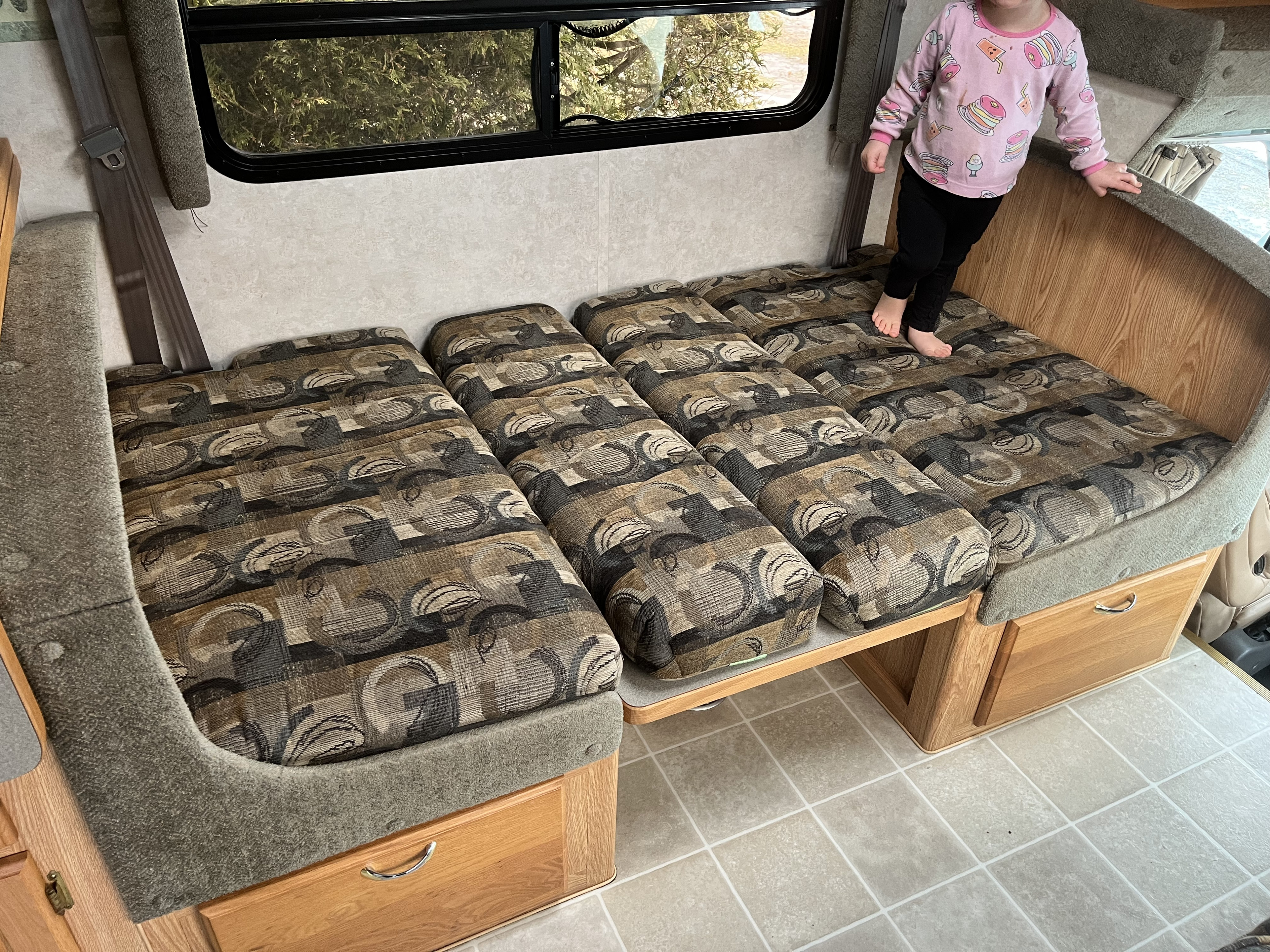 Cozy RV rental dinette with a child playing on the patterned cushions. Features wood storage, a window, and a seatbelt.. Bigfoot RV Bigfoot RV Motorhome 2004