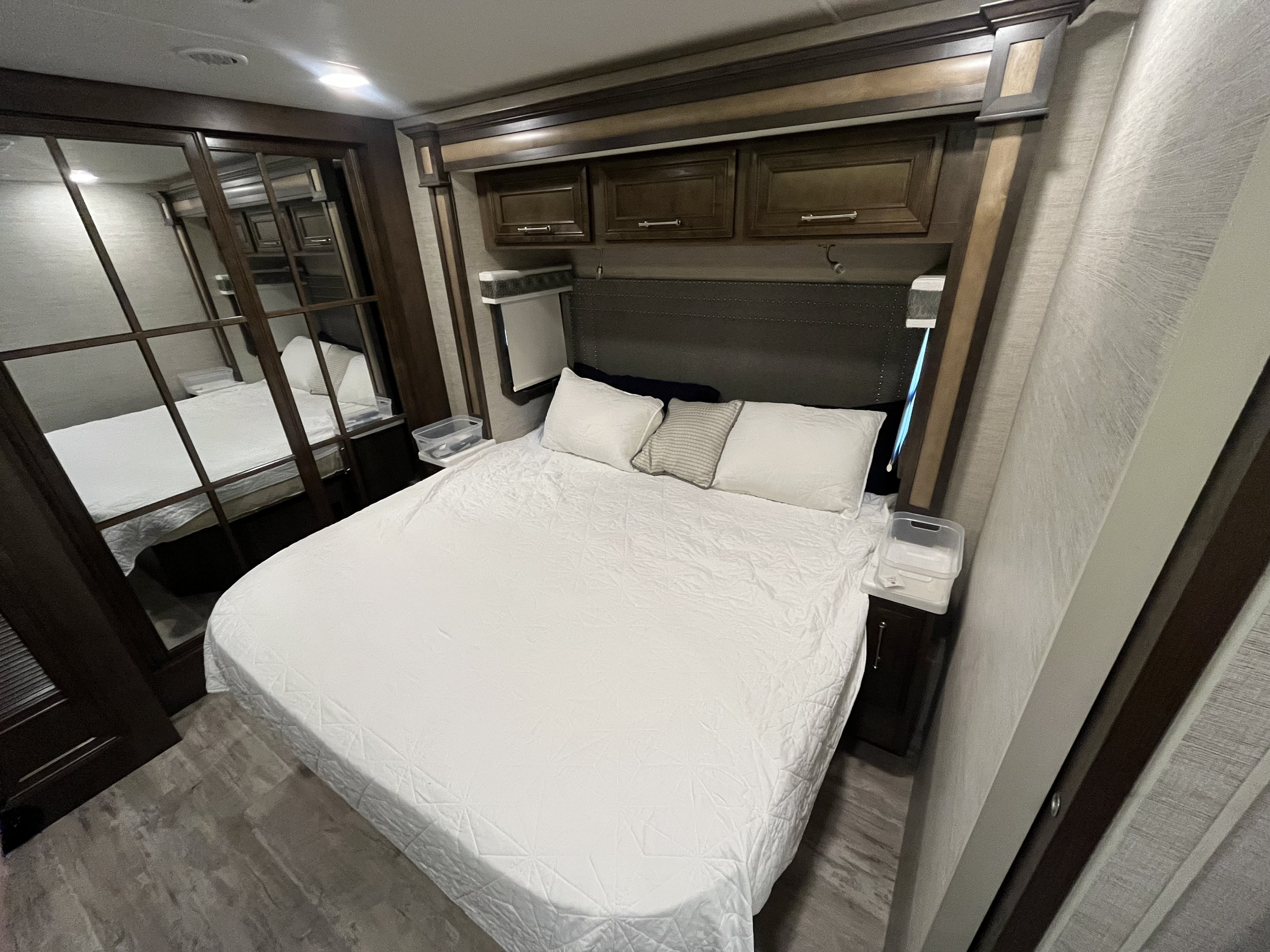 RV rental bedroom with white bed, dark headboard, mirrored closet, and overhead storage cabinets, offering comfort on the road.. Entegra Coach Emblem 2022
