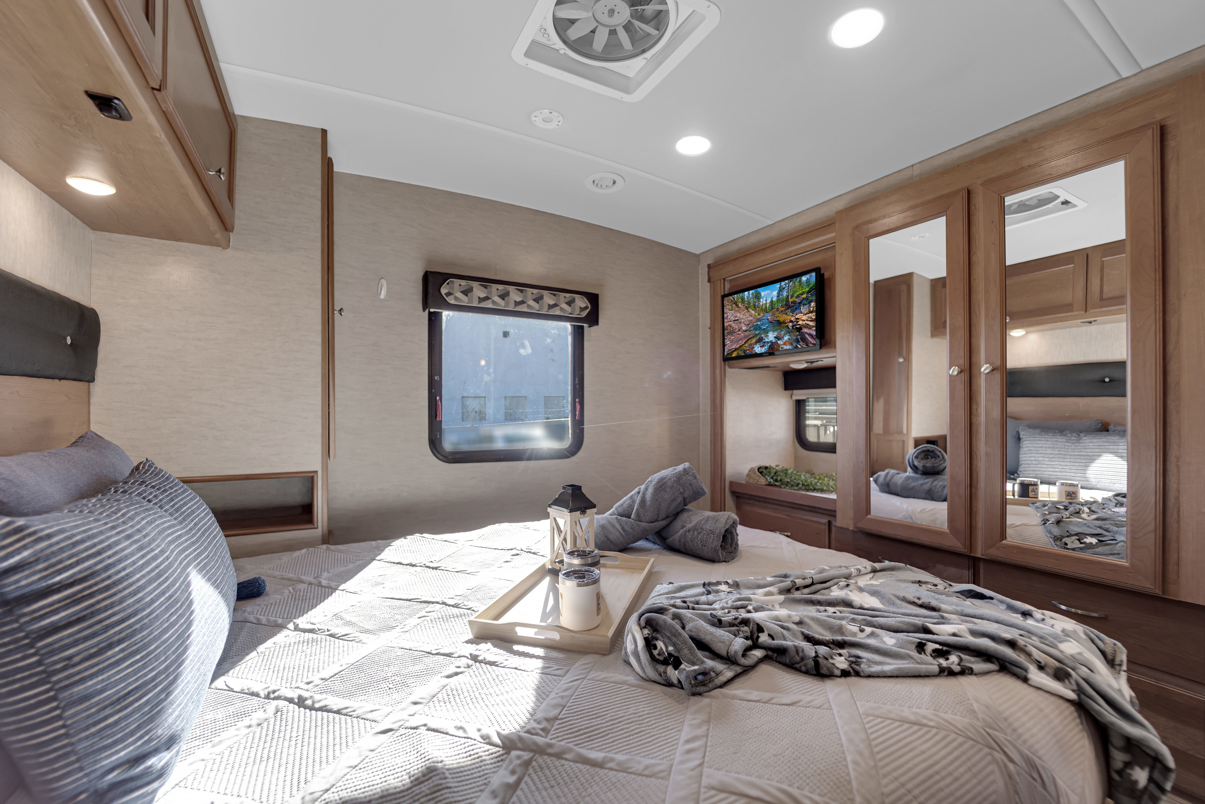 Bright RV rental bedroom: large bed with striped pillow & patterned blanket, window, overhead storage, TV, and mirrored closet.. Winnebago Vista 2017