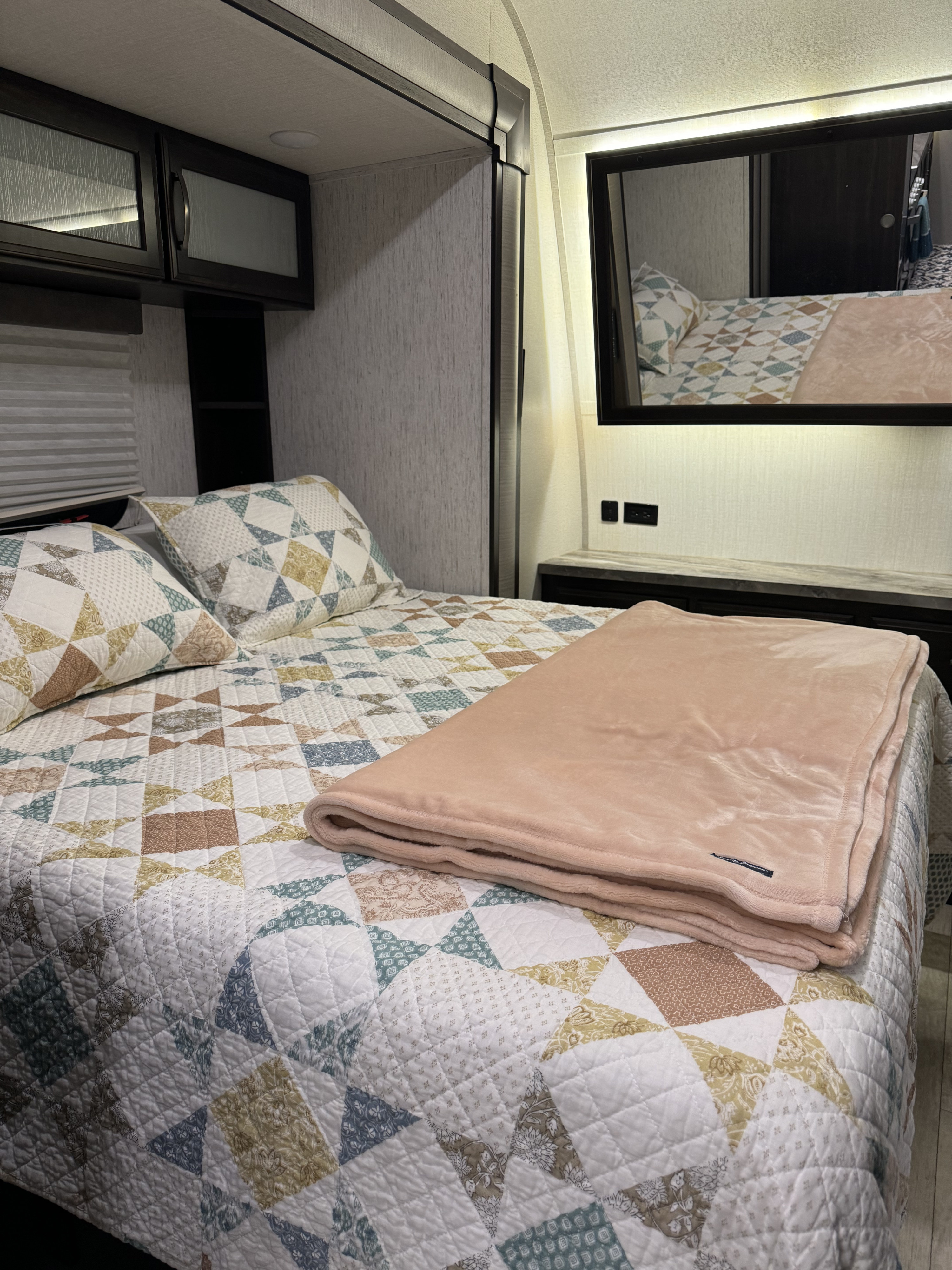 Cozy RV rental bedroom featuring a geometric quilt, soft pink blanket, overhead storage, and a mirrored vanity with ambient lighting.. Grand Design Momentum Toy Hauler 2020