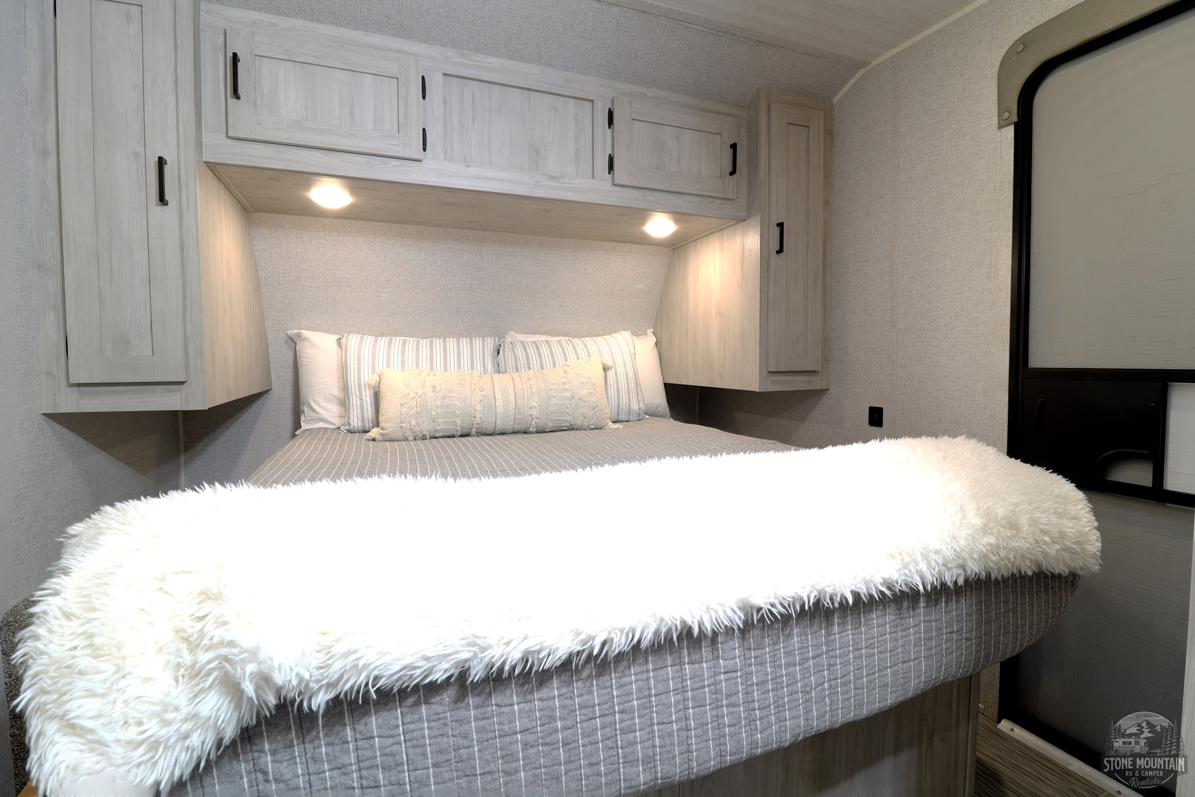 Comfortable RV rental bedroom with light wood overhead cabinets, soft textured walls, and a cozy bed with a fluffy white blanket.. East to West Silver Lake 2023
