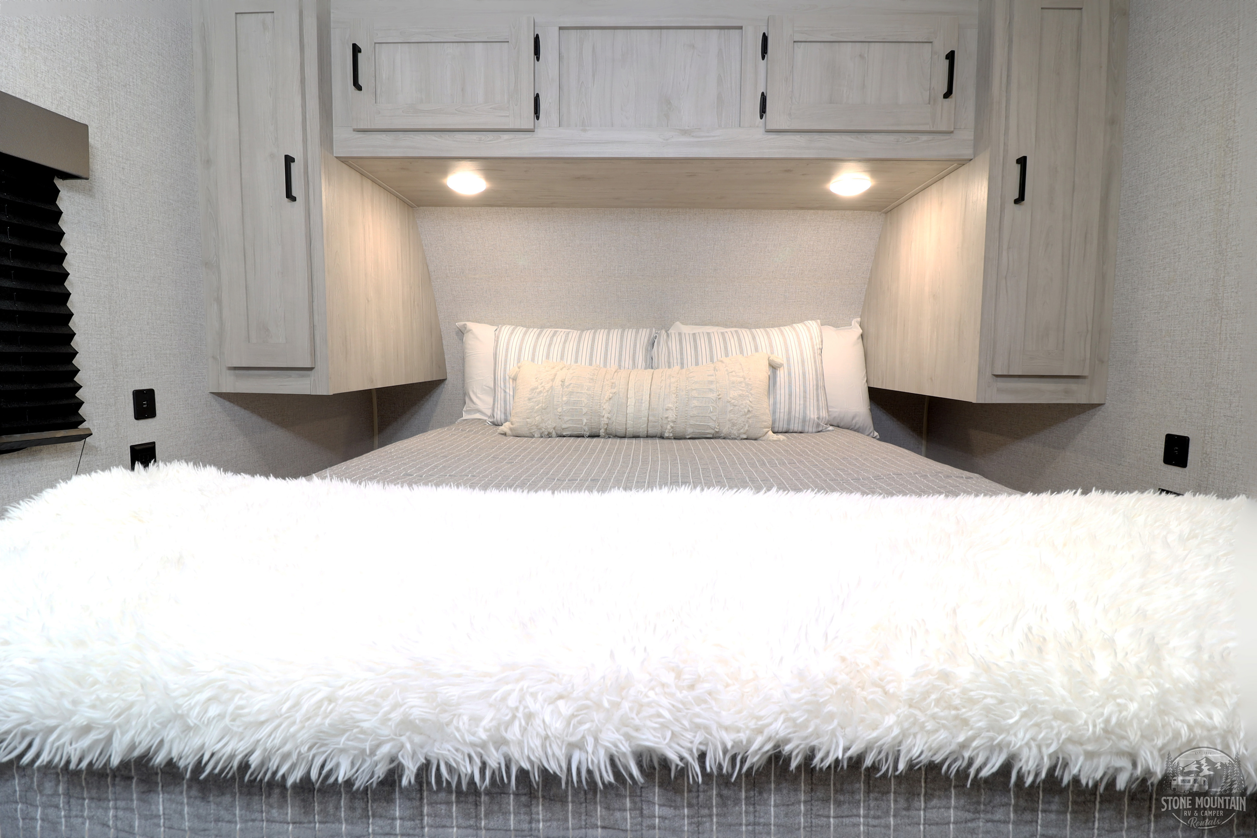 Experience comfort in this RV rental bedroom, featuring a cozy bed with a fluffy blanket, modern overhead cabinets, and soft lighting.. East to West Silver Lake 2023