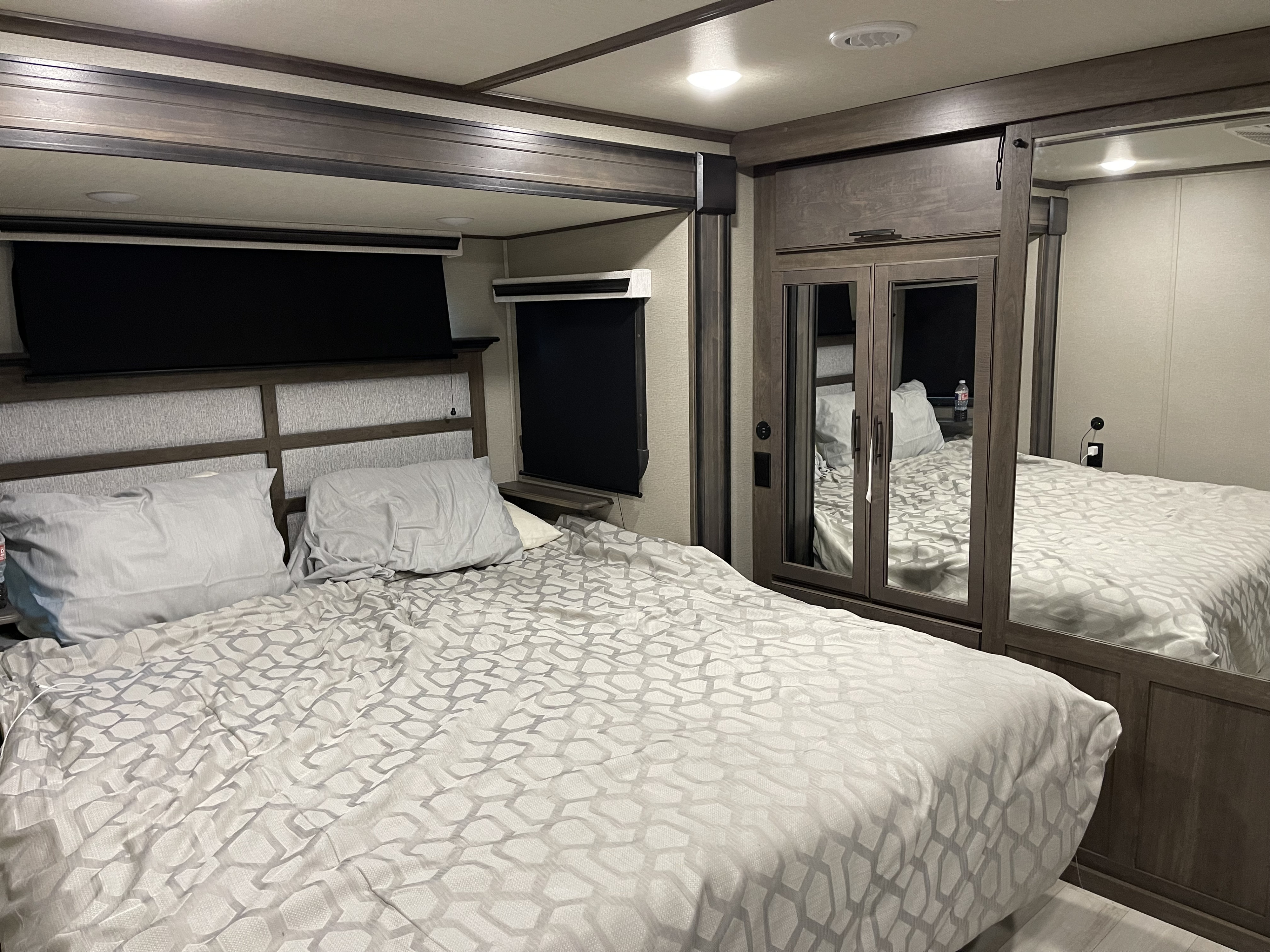 Cozy RV rental bedroom featuring a patterned bedspread, dark wood accents, black out shades, and a mirrored closet.. Grand Design Solitude 2022