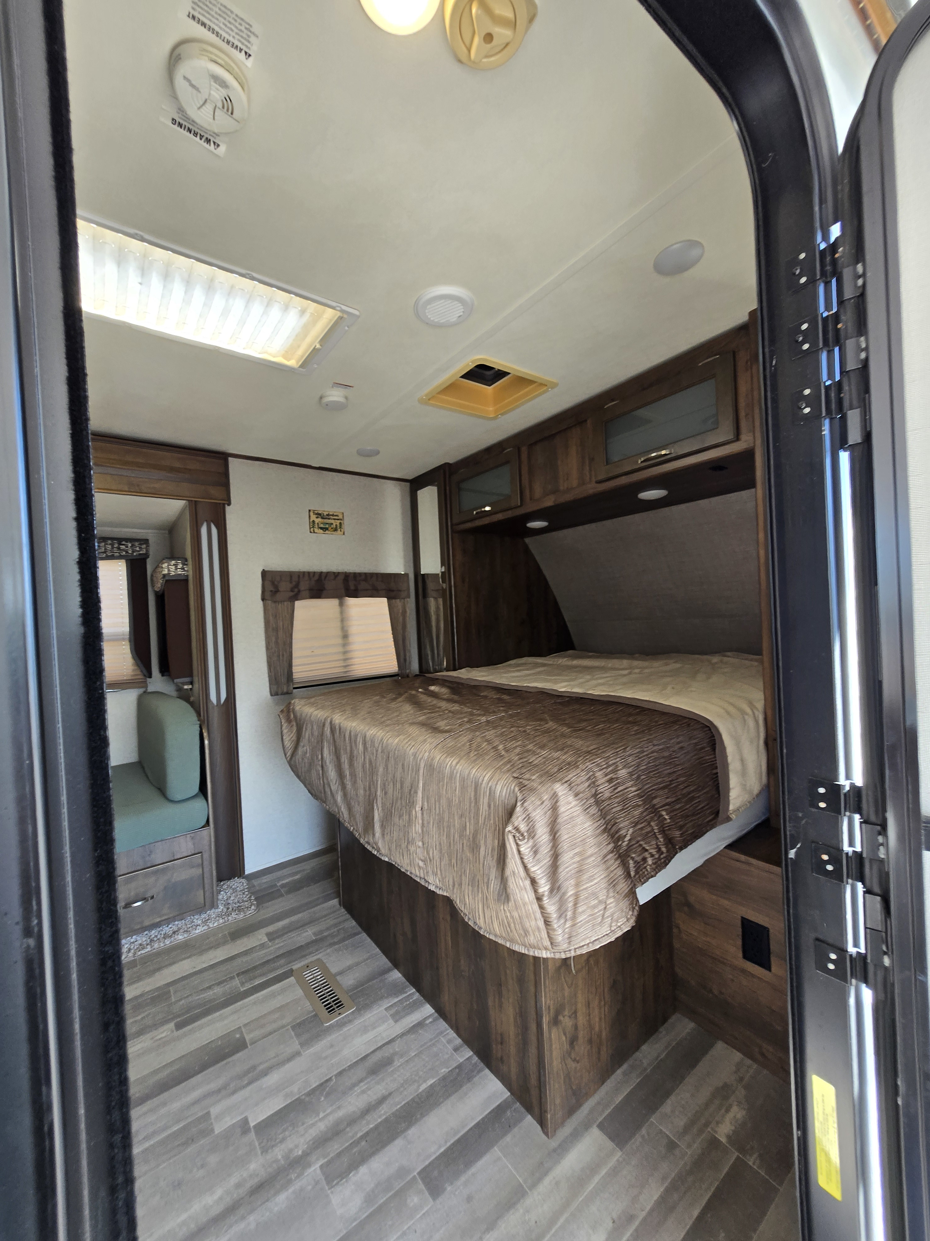 RV rental interior: A bedroom featuring a large bed with a brown comforter, overhead cabinets, wood-look flooring, and a side seating area.. Coachmen 2133RB 2019