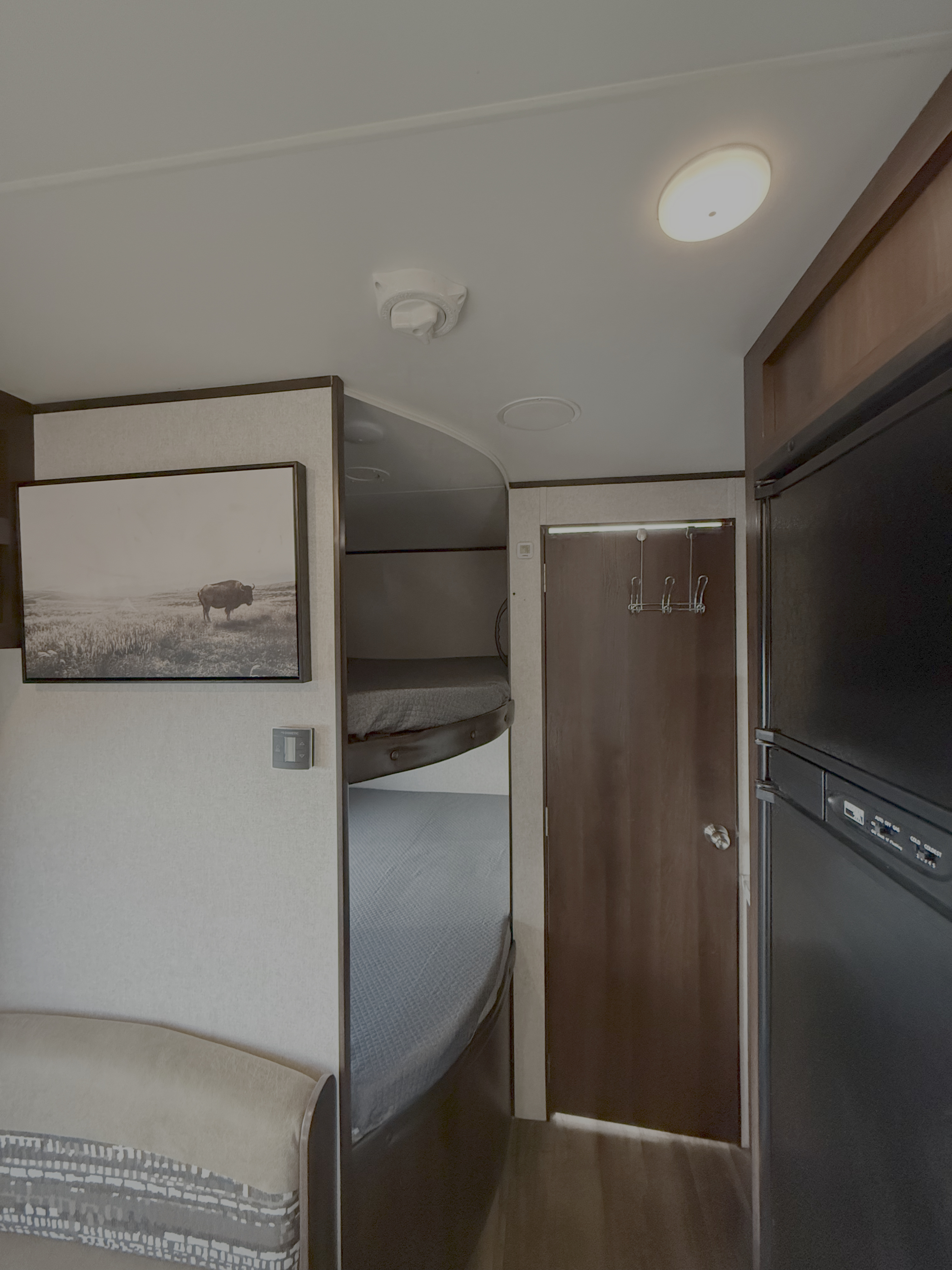 Bright RV rental interior with bunk beds, a dark wood door, and refrigerator. Wall art and thermostat provide a homelike touch.. Jayco Jay Flight SLX 2019