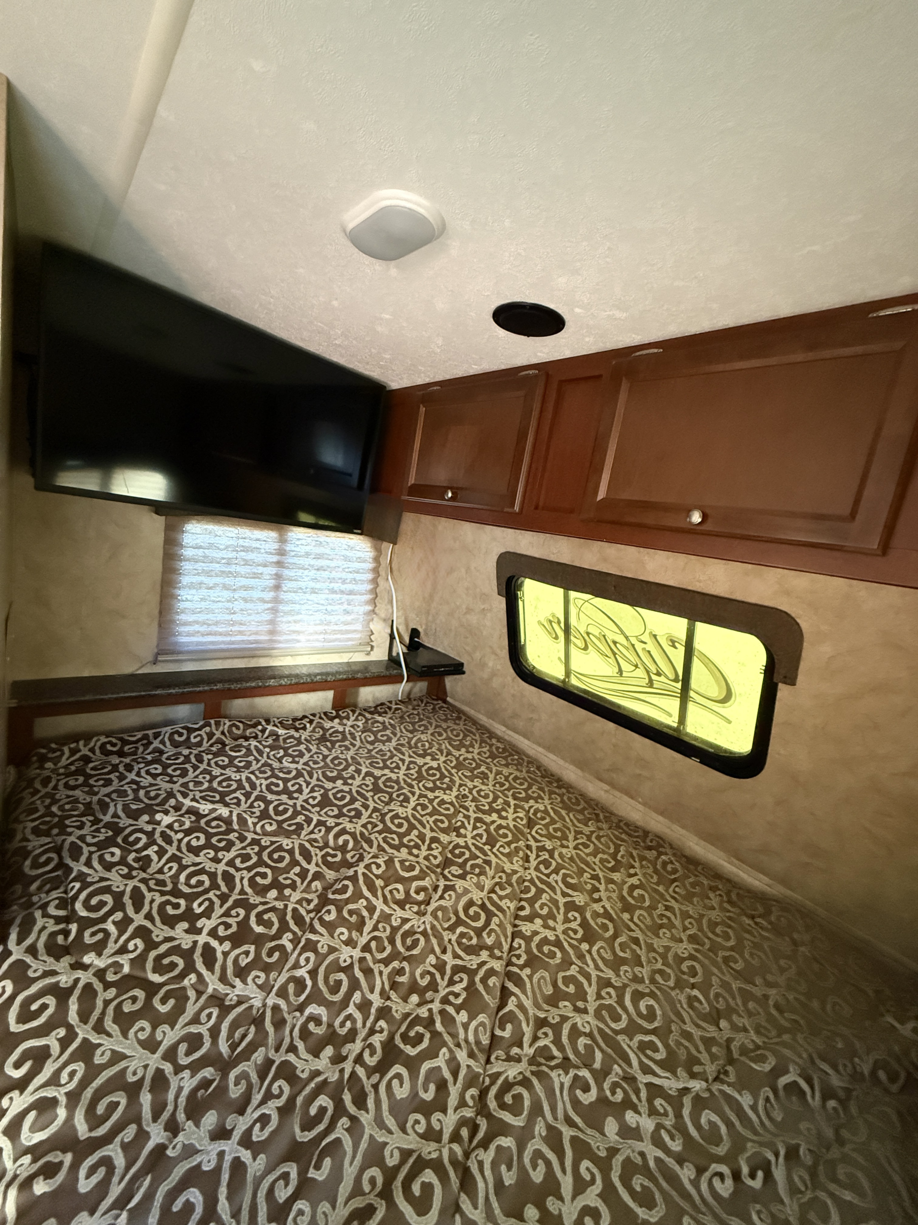 Cozy RV rental bedroom with a brown & white patterned bed, wall-mounted TV, overhead cabinets, and two windows.. Coachmen Clipper 2016