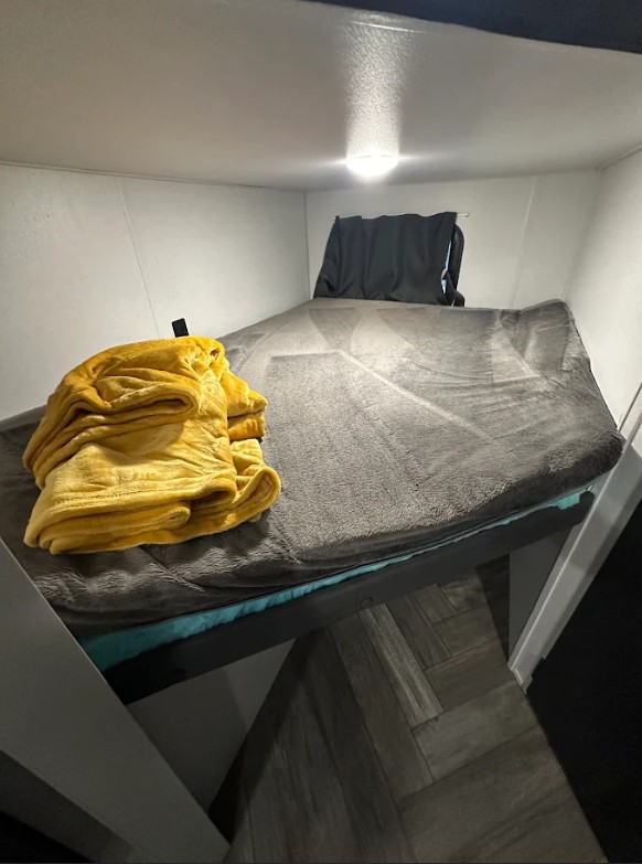 Inside an RV rental: a compact bunk bed with a soft gray blanket, folded yellow blanket, and ceiling light.. Forest River Wildwood FSX 2021