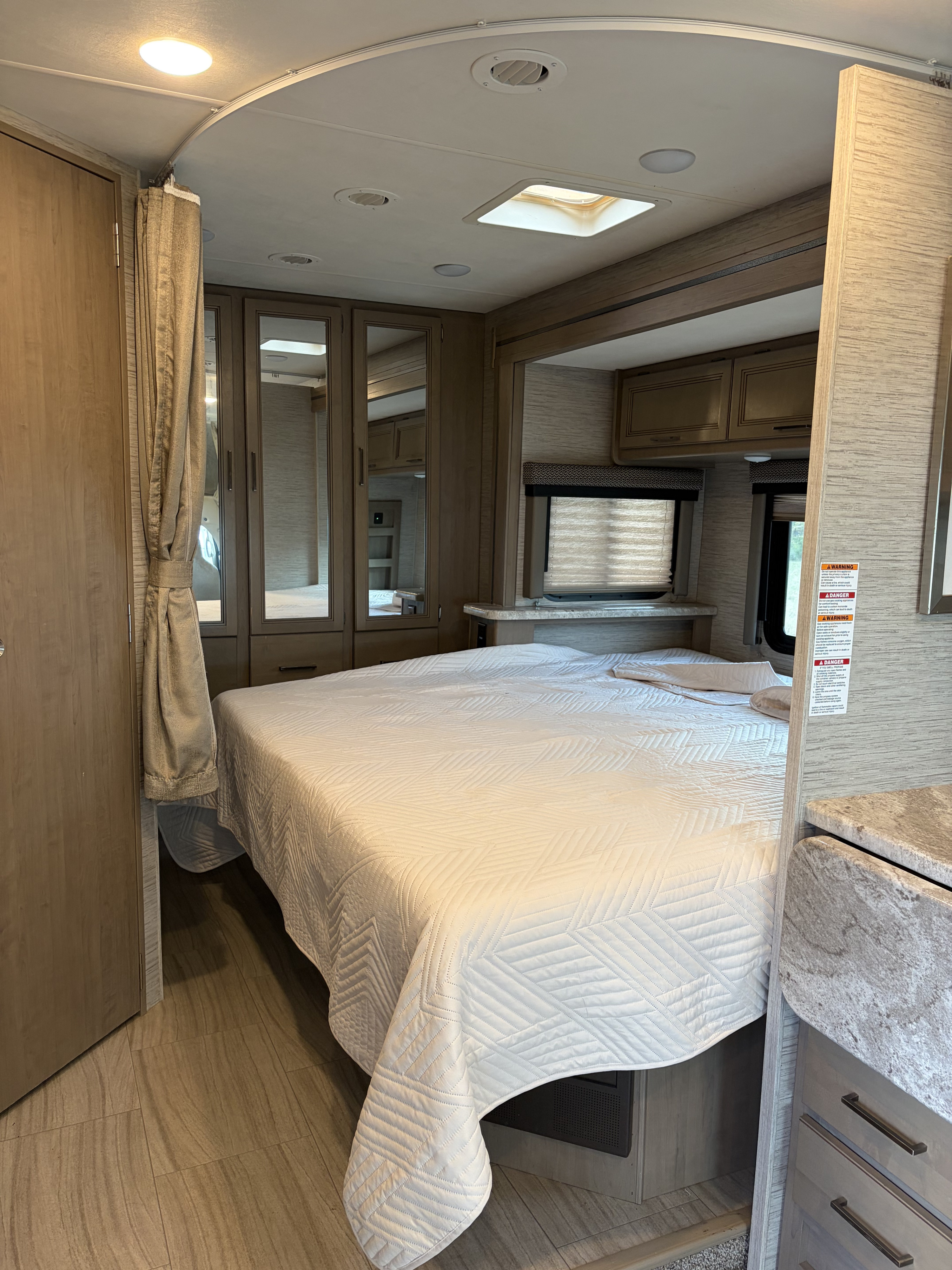 Experience comfort in this RV rental bedroom, featuring a white quilted bed, mirrored storage cabinets, and natural light from windows.. Thor Daybreak 2021