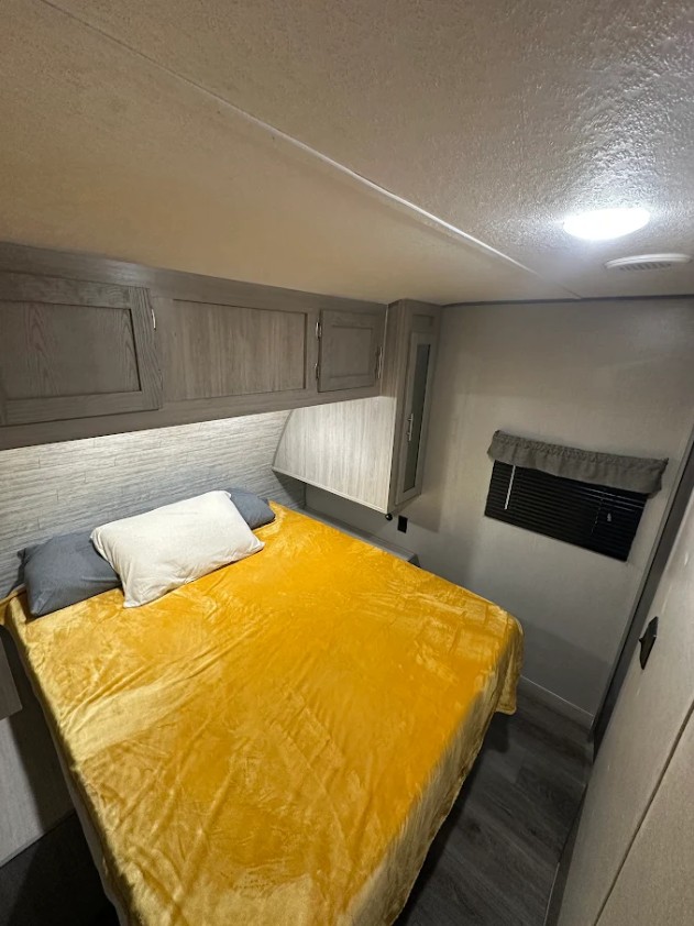 This bright and cozy RV rental bedroom features a yellow bedspread, overhead storage, and a window with blinds.. Forest River Coachmen Catalina Legacy 2022