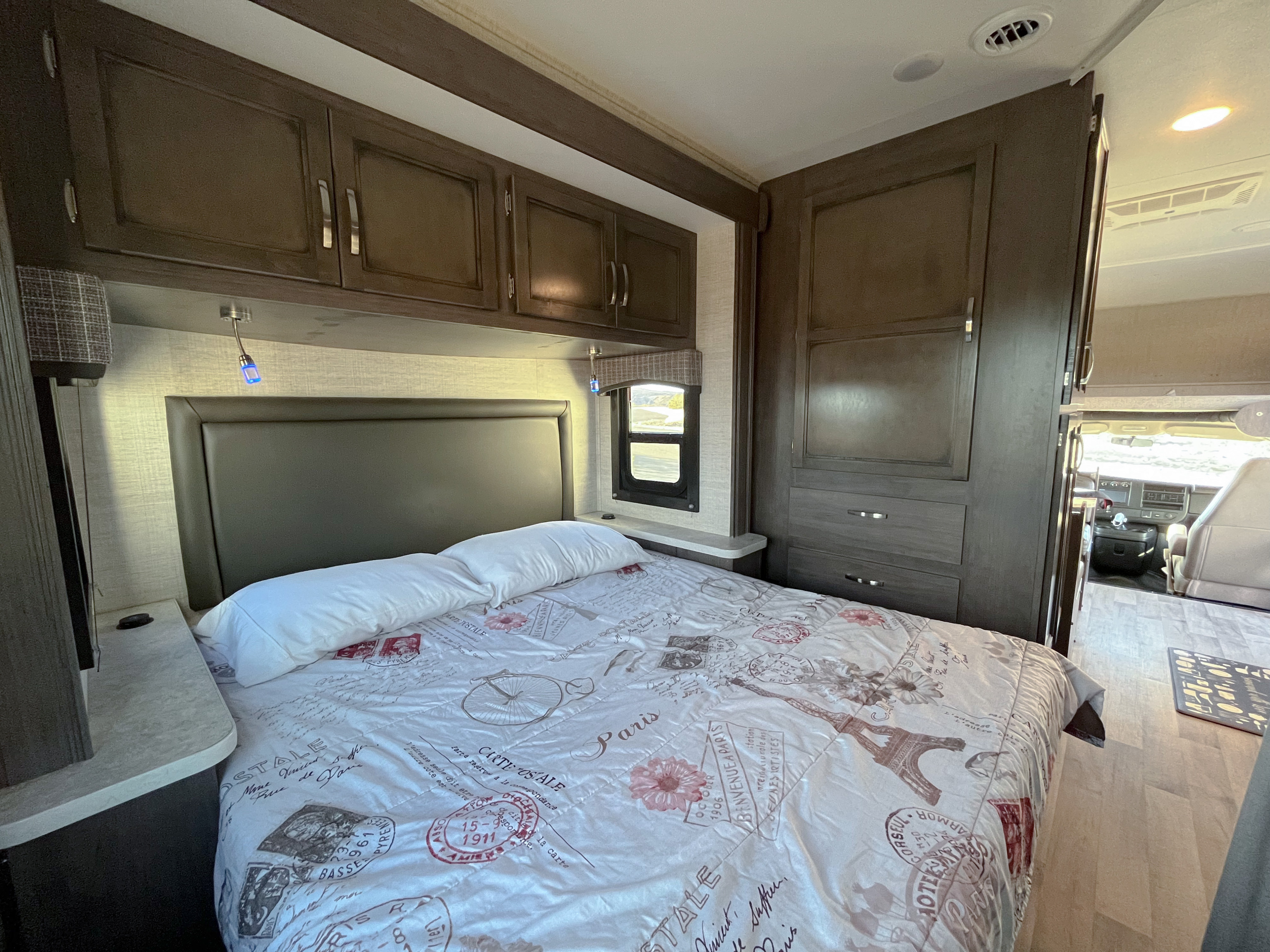 Compact RV rental bedroom featuring Paris-themed bedding, overhead storage, reading light, and a view into the cockpit.. Entegra Coach Odyssey 2023