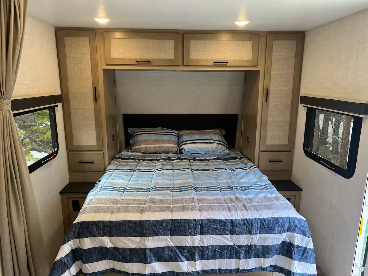 Cozy rv rental bedroom showcasing a bed with striped bedding, practical built-in storage, and windows with scenic views.. Ember E 2024