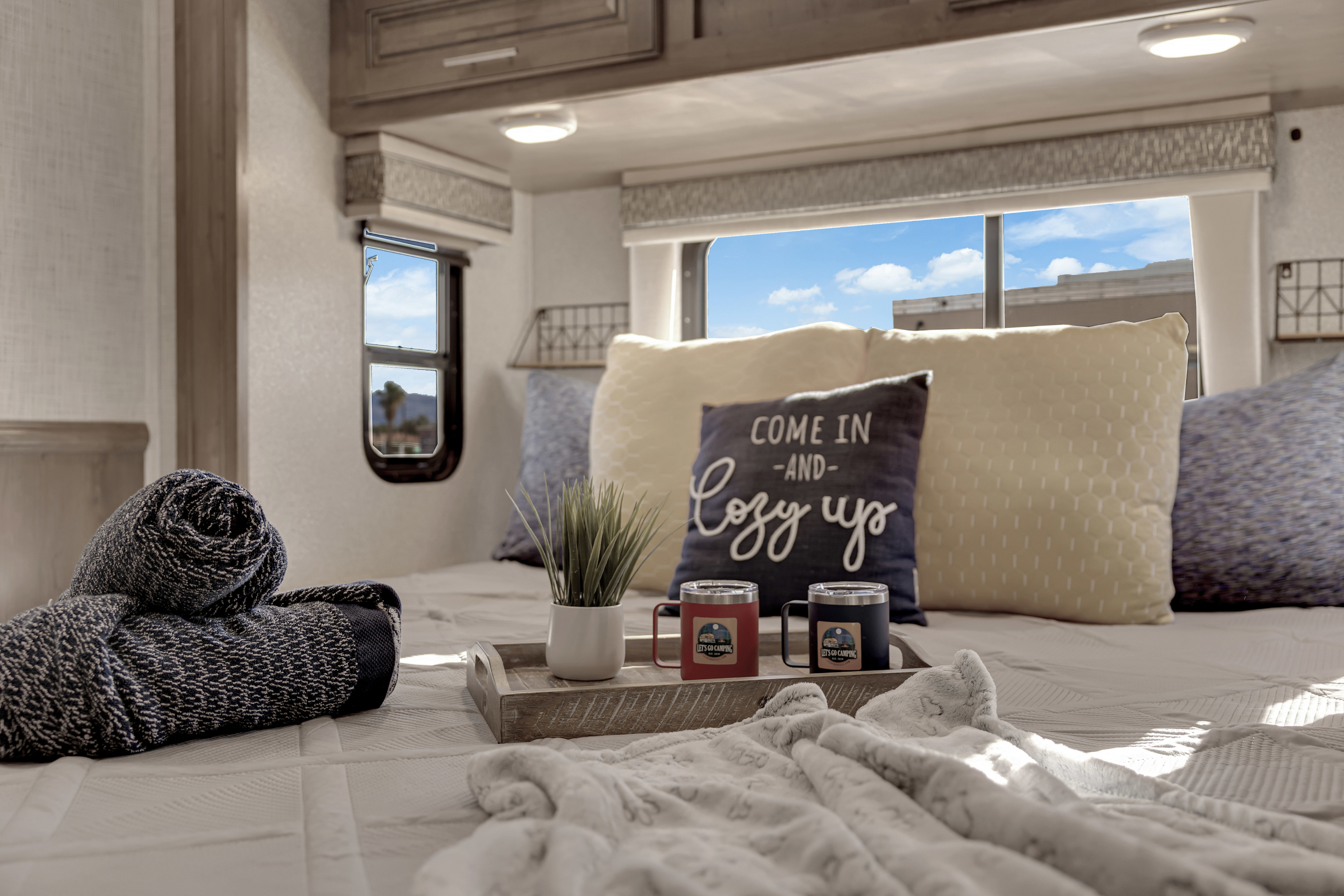 Cozy RV rental bedroom with 'Come in and Cozy up' pillow, blankets, plant, and 'Camping' mugs on a tray by sunny windows.. Forest River Georgetown 2019
