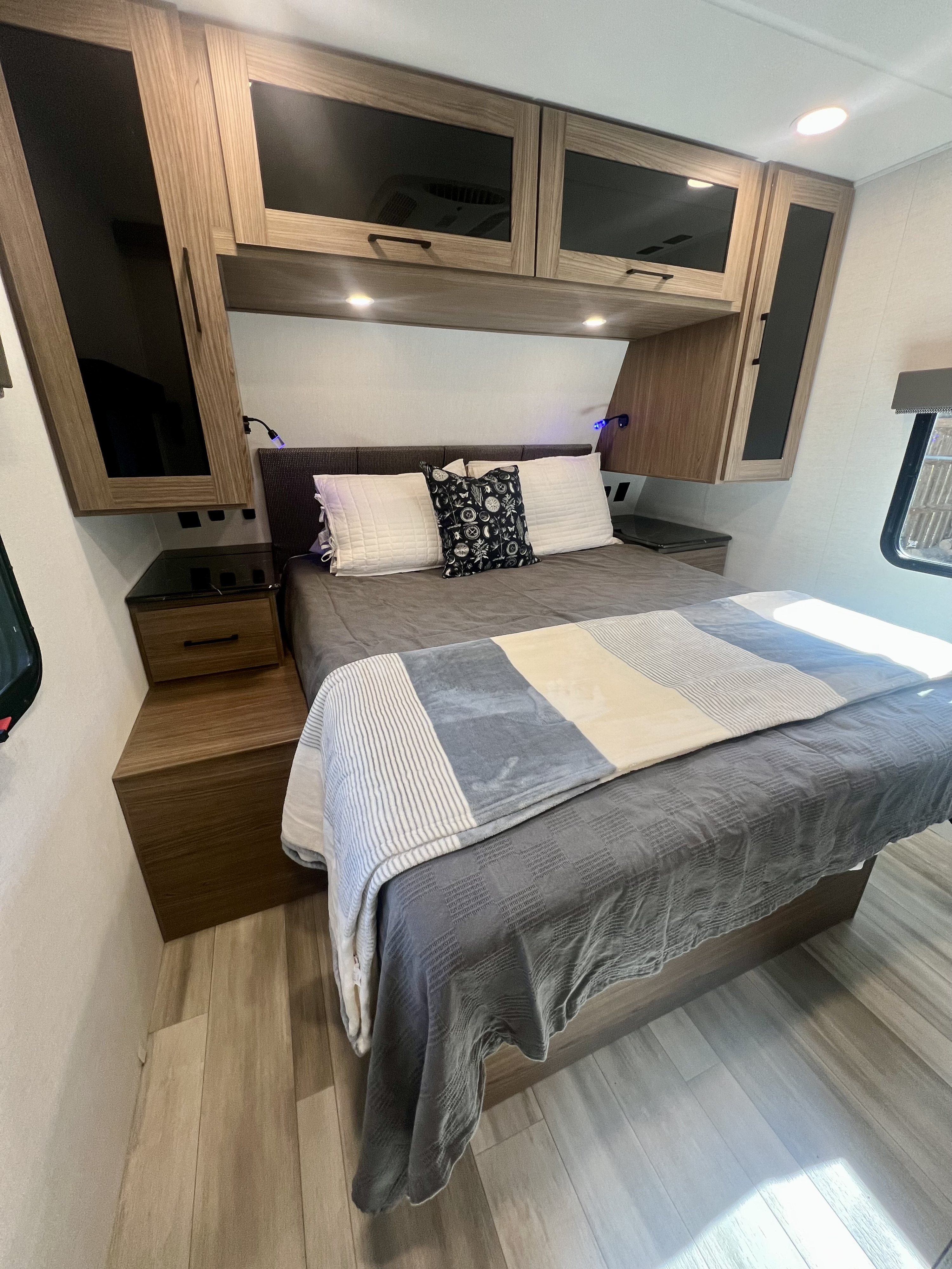 RV rental bedroom: queen bed with grey bedding, overhead wood cabinets, built-in nightstands & modern lighting for a relaxing stay.. Alliance RV Delta 2025