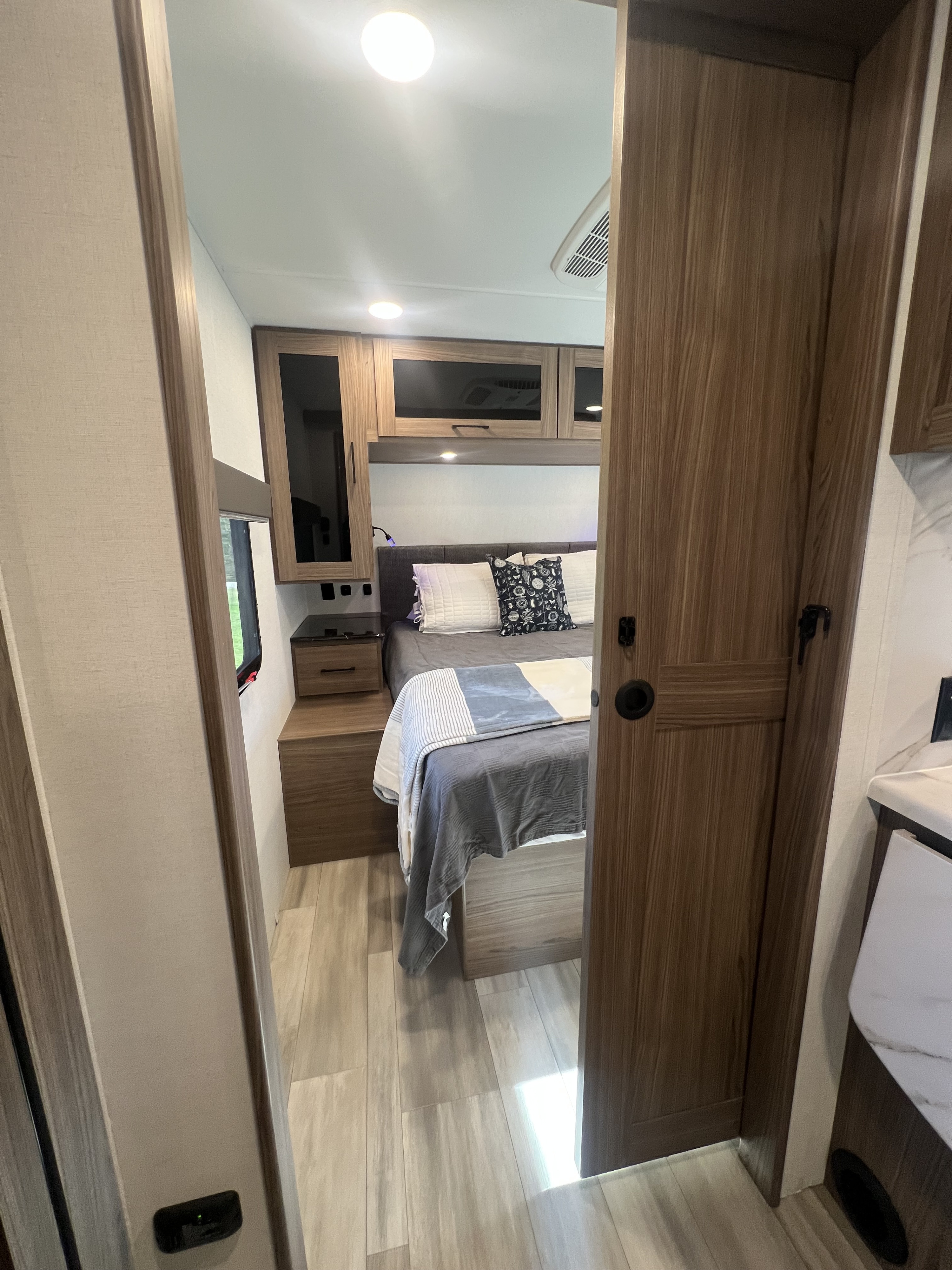 View into a comfortable RV rental bedroom, showing a bed, built-in nightstands, overhead cabinets, and wood-look flooring.. Alliance RV Delta 2025