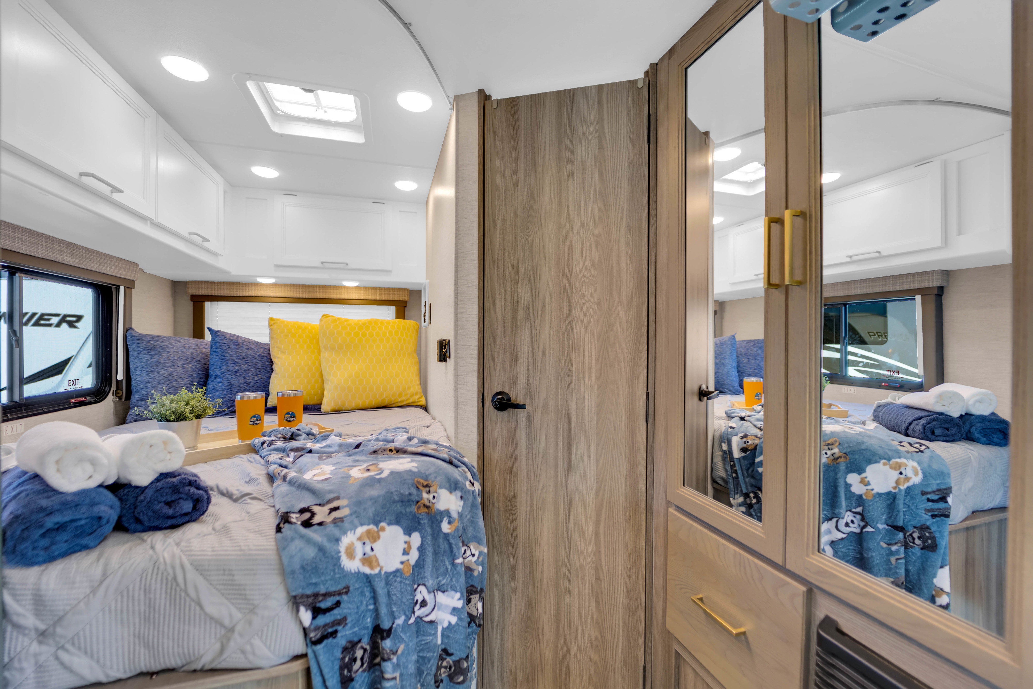 Cozy RV rental bedroom featuring a comfy bed with dog-patterned blanket, blue & yellow pillows, towels, & overhead storage.. Thor Eddie Bauer 2025