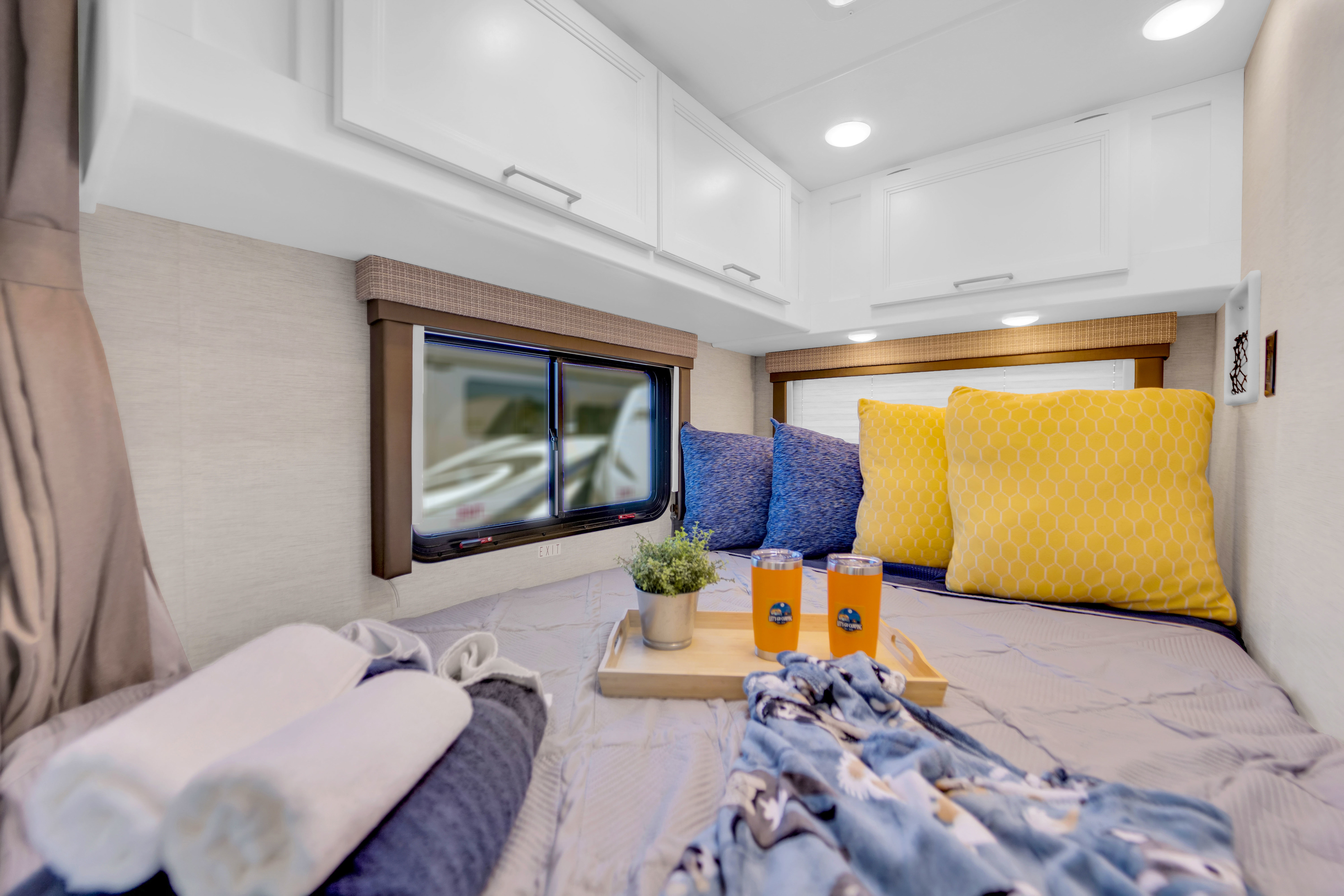 Cozy RV rental bedroom with a bed, blue and yellow pillows, overhead cabinets, and a tray with tumblers. Perfect for travel.. Thor Eddie Bauer 2025