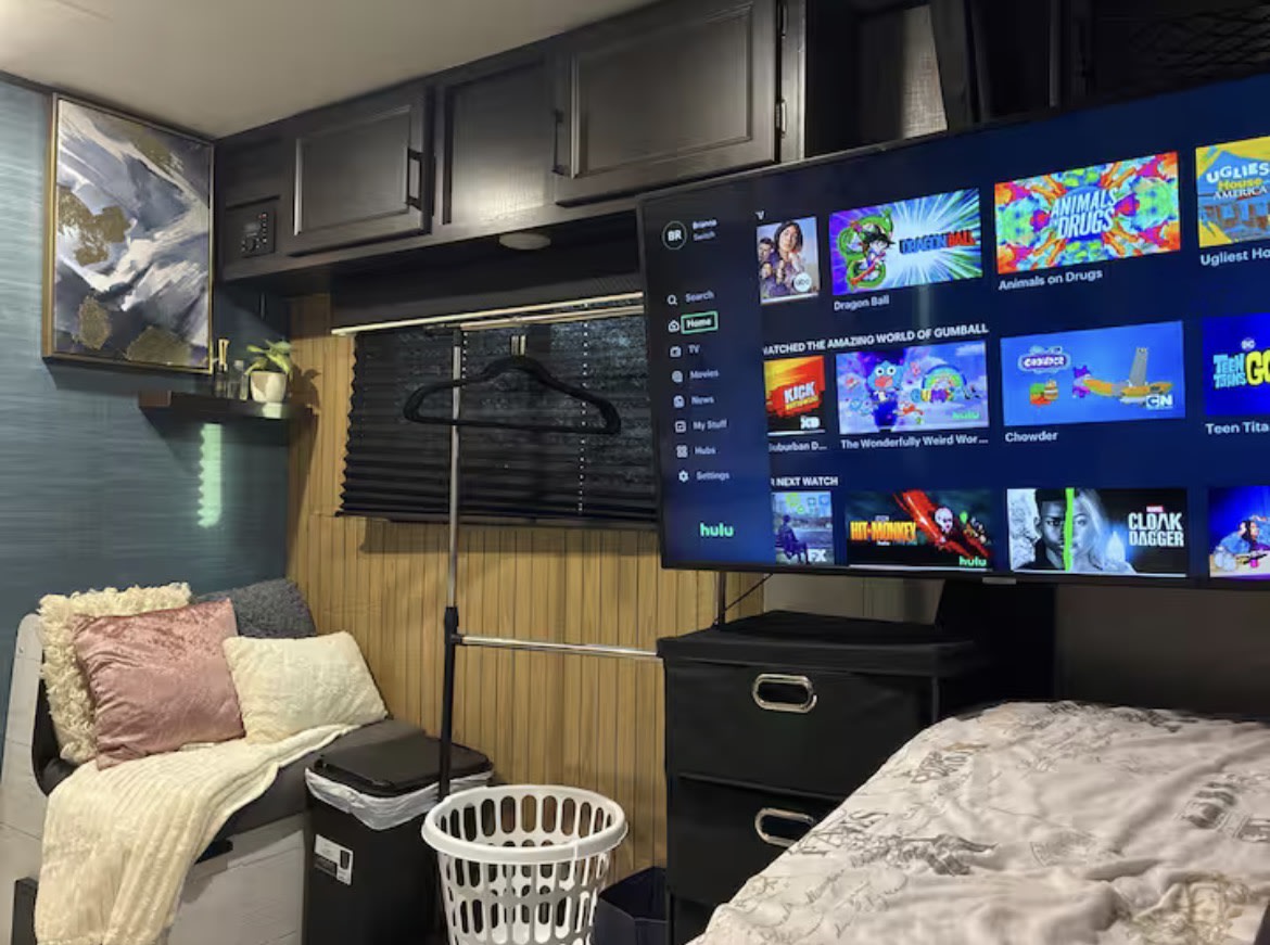 Cozy RV rental interior featuring a TV displaying Hulu, a comfy bench, dark storage cabinets, and a clothes rack.. Jayco Jay Flight SLX 2023