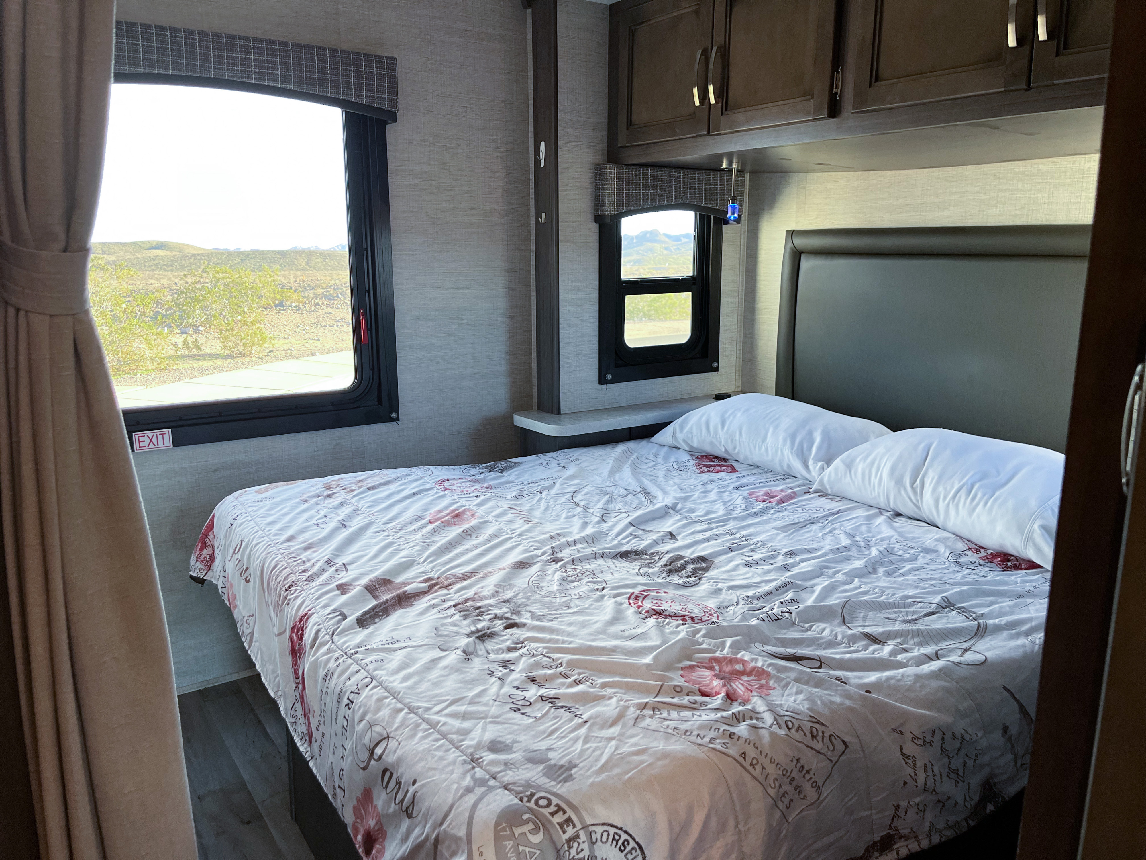 Cozy rv rental bedroom with a Paris-themed bed, overhead cabinets, and a desert view from the window.. Entegra Coach Odyssey 2023