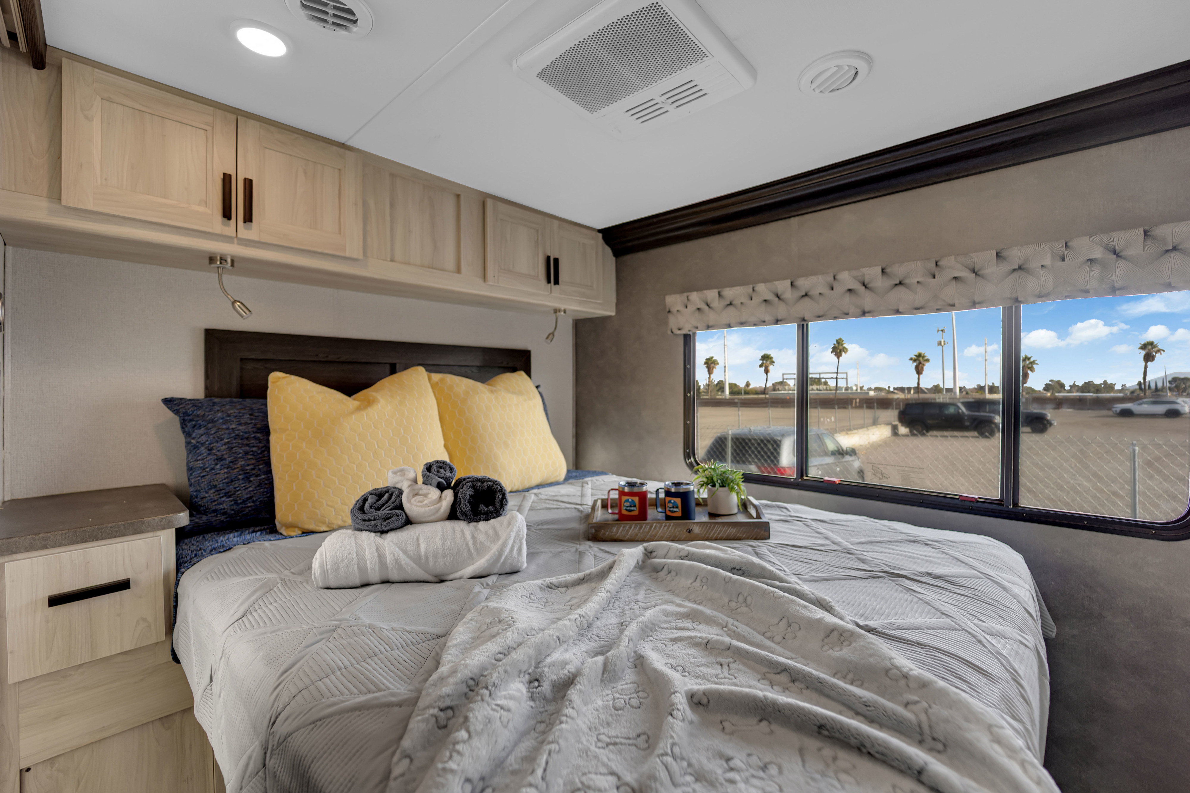 Relaxing RV rental bedroom: light wood cabinets, reading lights, a bed with yellow pillows, and a window showing palm trees.. Forest River Sunseeker 2021