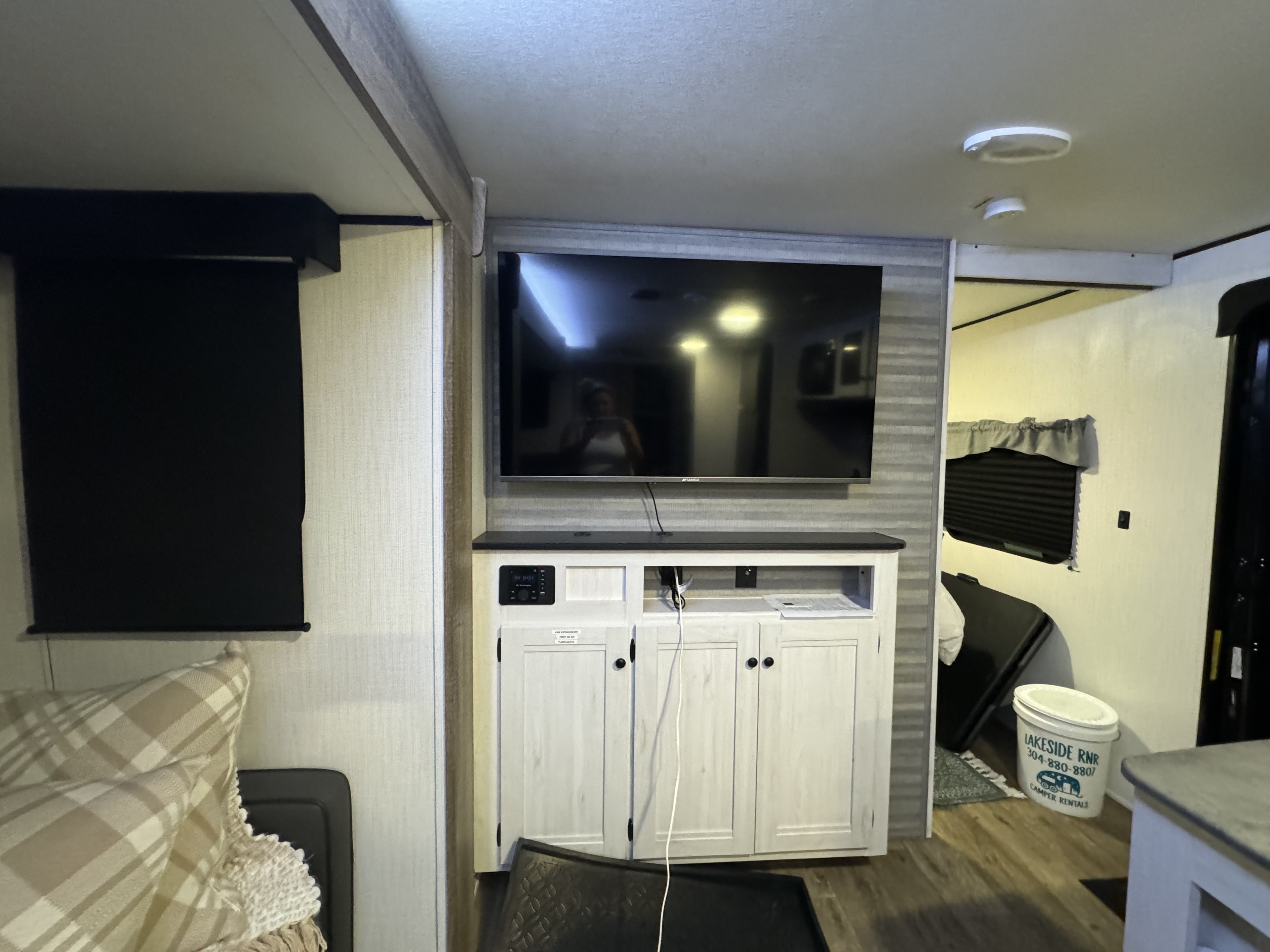 RV rental interior featuring a TV above a media console, a drawn roller blind, and a "Lakeside RNR Camper Rentals" bucket.. Heartland RVs Trail Runner 2024