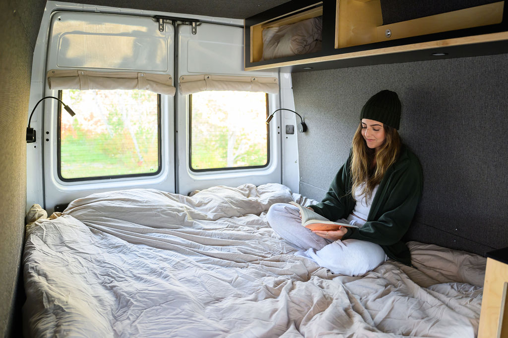 Relaxing in an RV rental: woman reads on a cozy campervan bed with windows showing nature. Ideal for travel.. Dodge Sprinter 2003