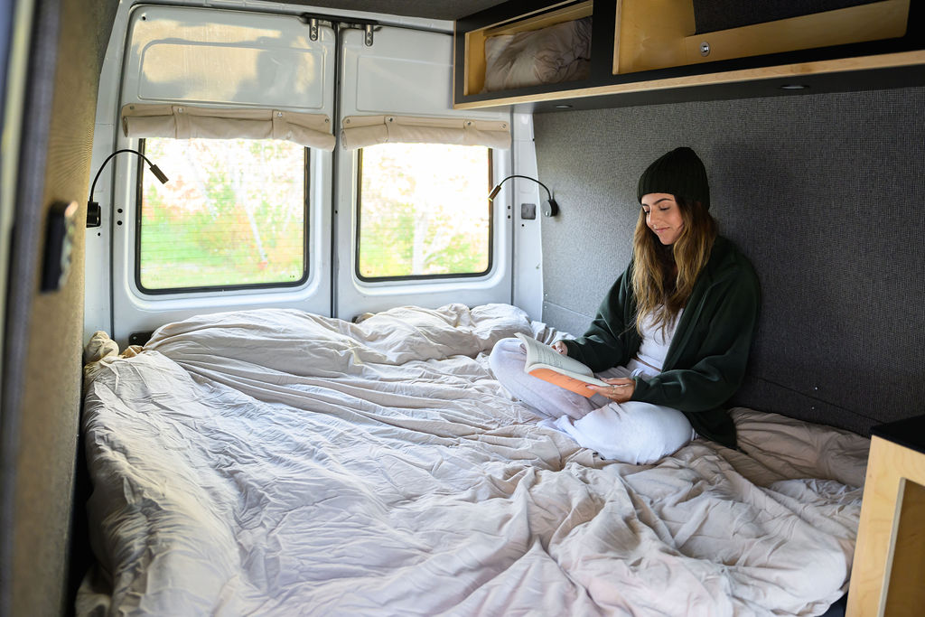 Cozy RV rental living: A woman reads on a comfy bed in her camper van, windows showing lush nature outside.. Dodge Sprinter 2005