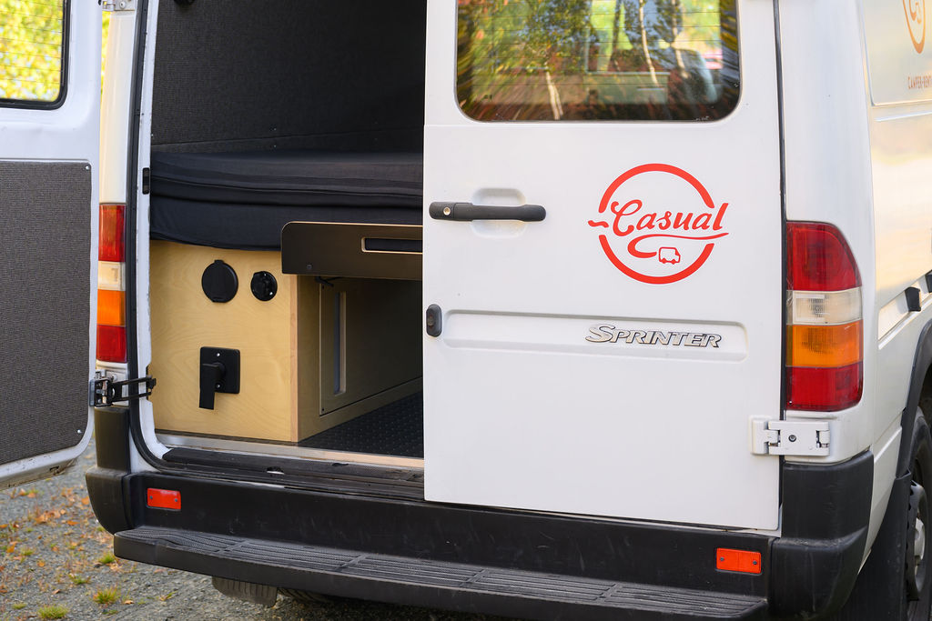 Open rear of a white Sprinter rv rental van, showing custom interior with bed, storage, power outlets, and "Casual" logo.. Dodge Sprinter 2005