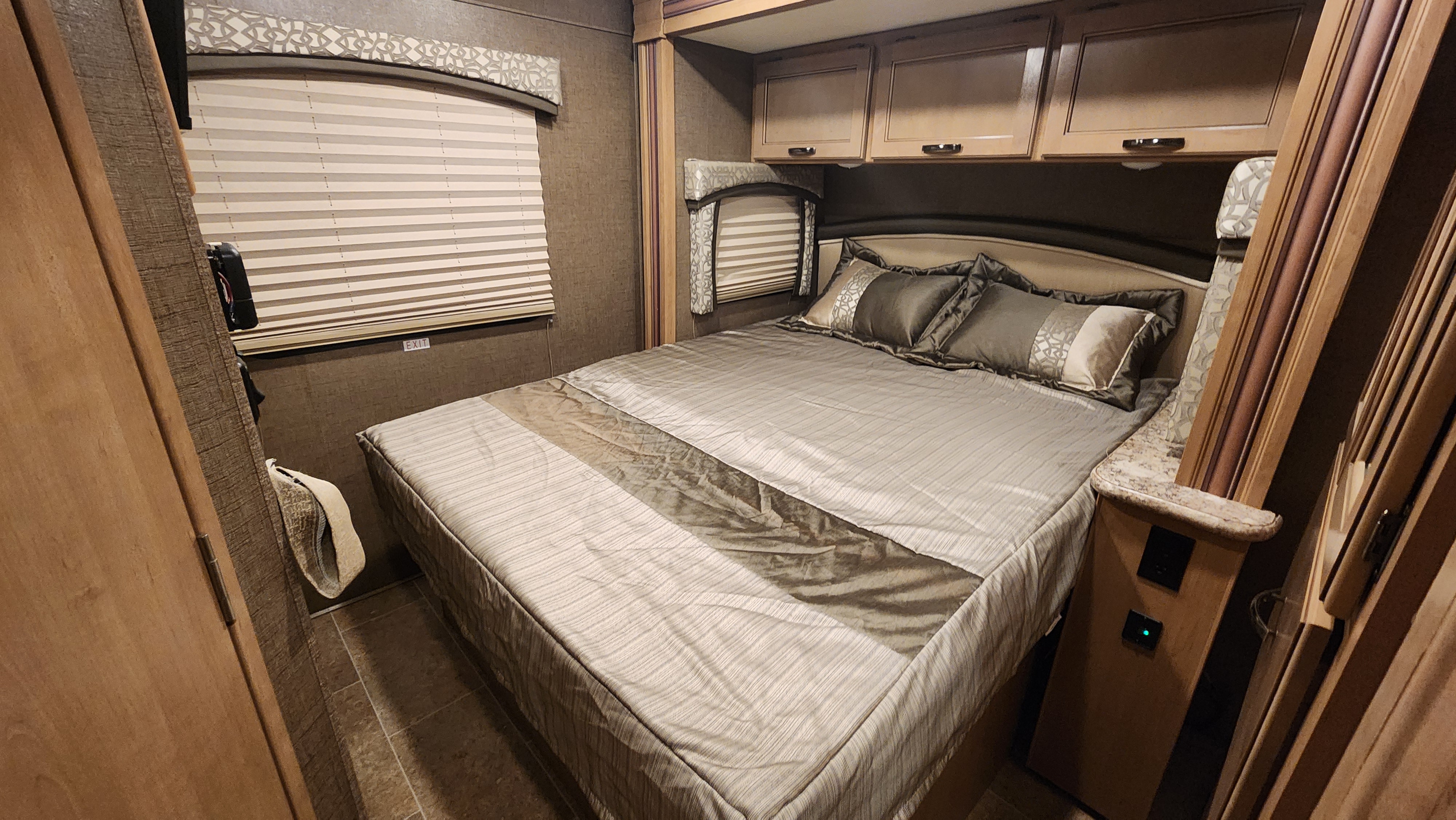 RV rental bedroom view: neutral-toned bed with pillows, wooden overhead cabinets, and pleated window blinds. Cozy and functional.. Thor Chateau 2019