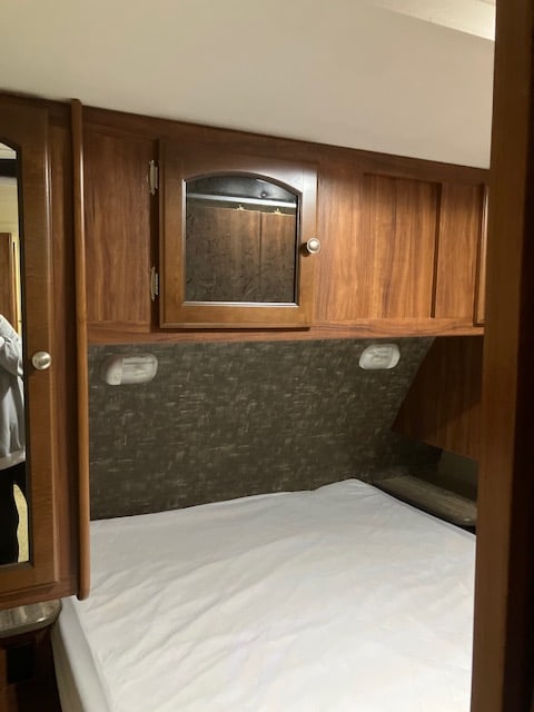 Inside an RV rental: a compact bedroom with a white-sheeted bed, wood cabinets, mirrored storage, and wall sconces.. Heartland RVs North Trail 2015