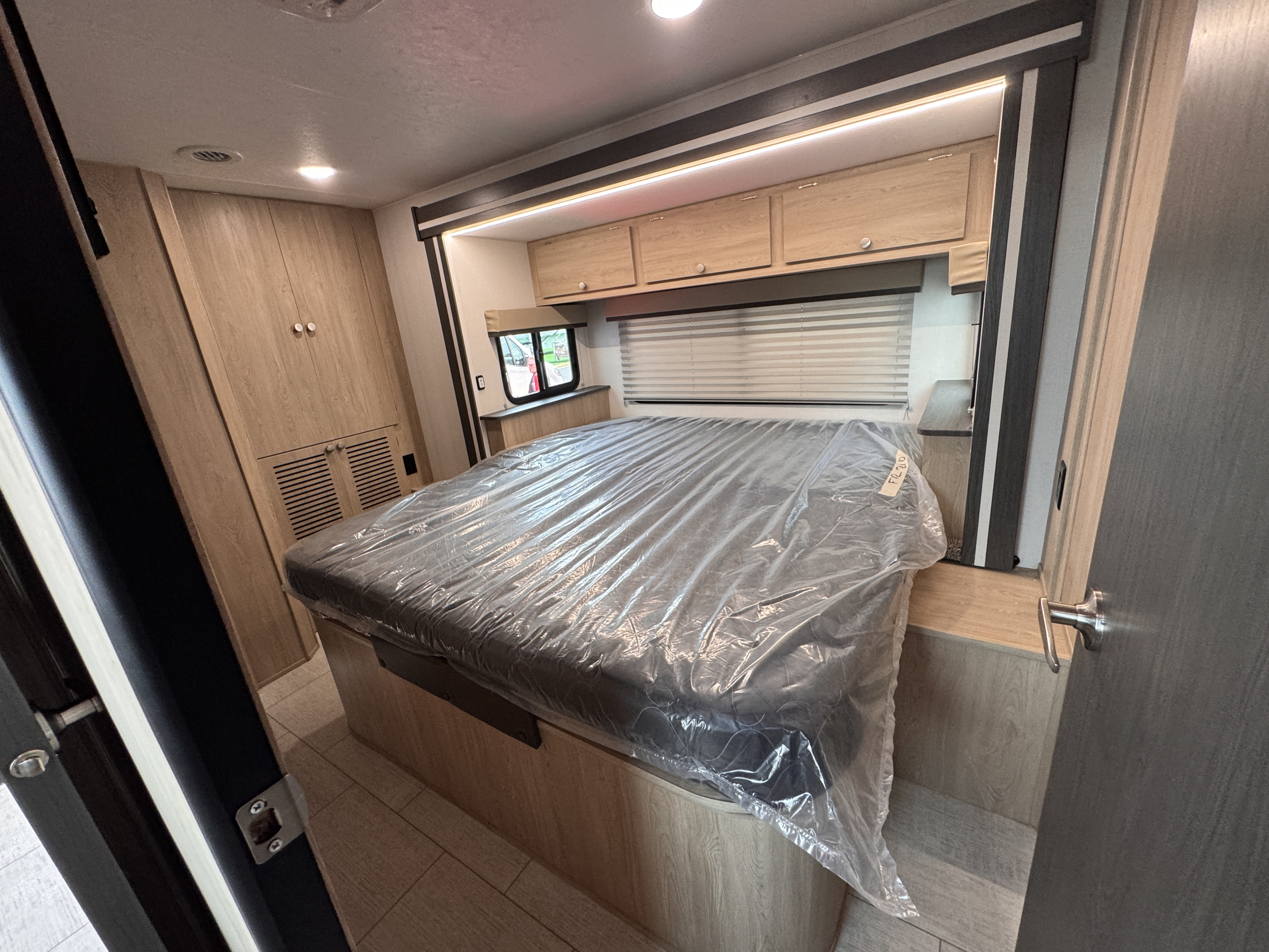 Bright RV rental bedroom featuring a bed covered in protective plastic, wood cabinets, and a window.. Forest River Coachmen Freelander 2026