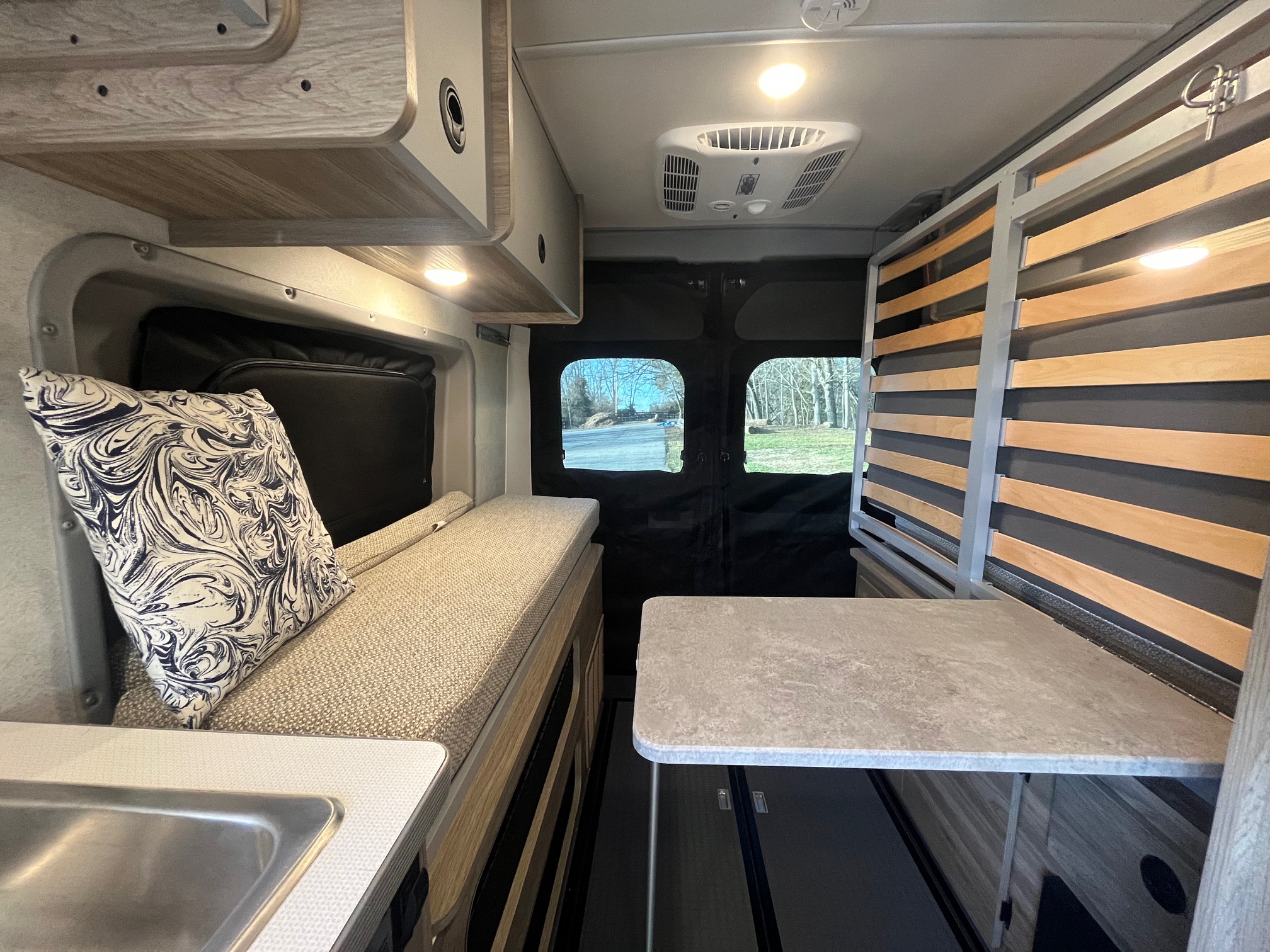 This rv rental interior has a cozy bench, sink, table, and slatted bed frame, with bright overhead lights and outdoor views.. Winnebago Solis 2025