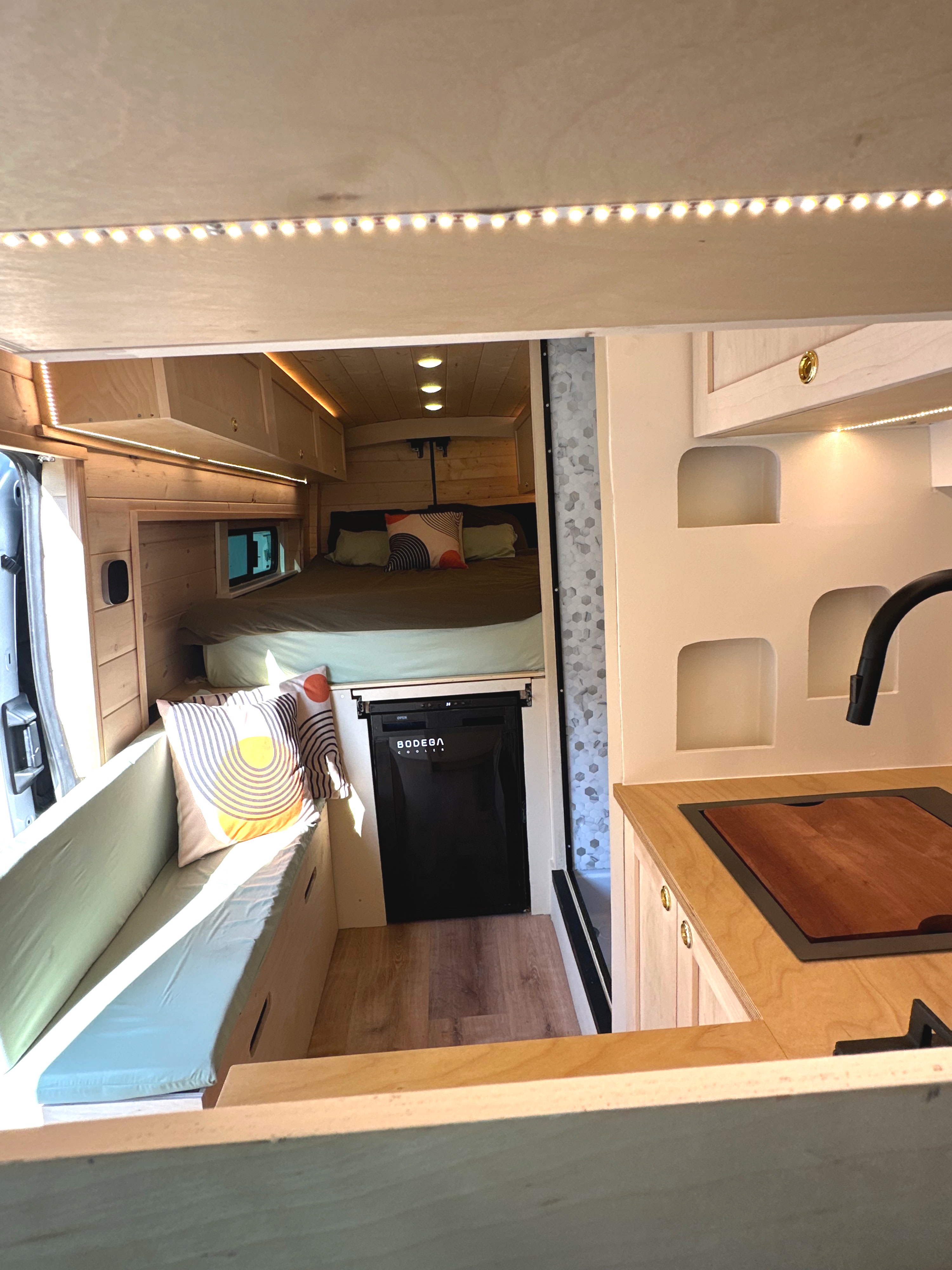This rv rental shows a cozy van interior with a bed, kitchen sink, Bodega cooler, and bench seating, lit by warm LED strips.. Panoramic RV Panoramic RV Class B 2023