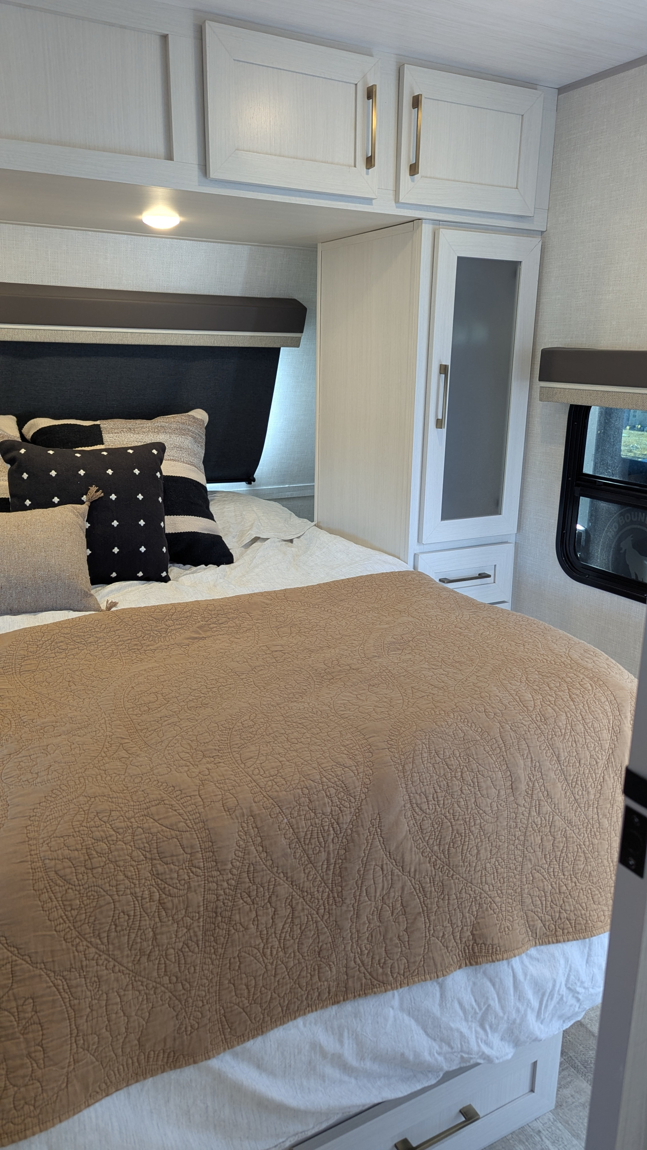Cozy RV rental bedroom featuring a bed with a quilted blanket, patterned pillows, overhead cabinets, and a window.. No Boundaries 20 2025