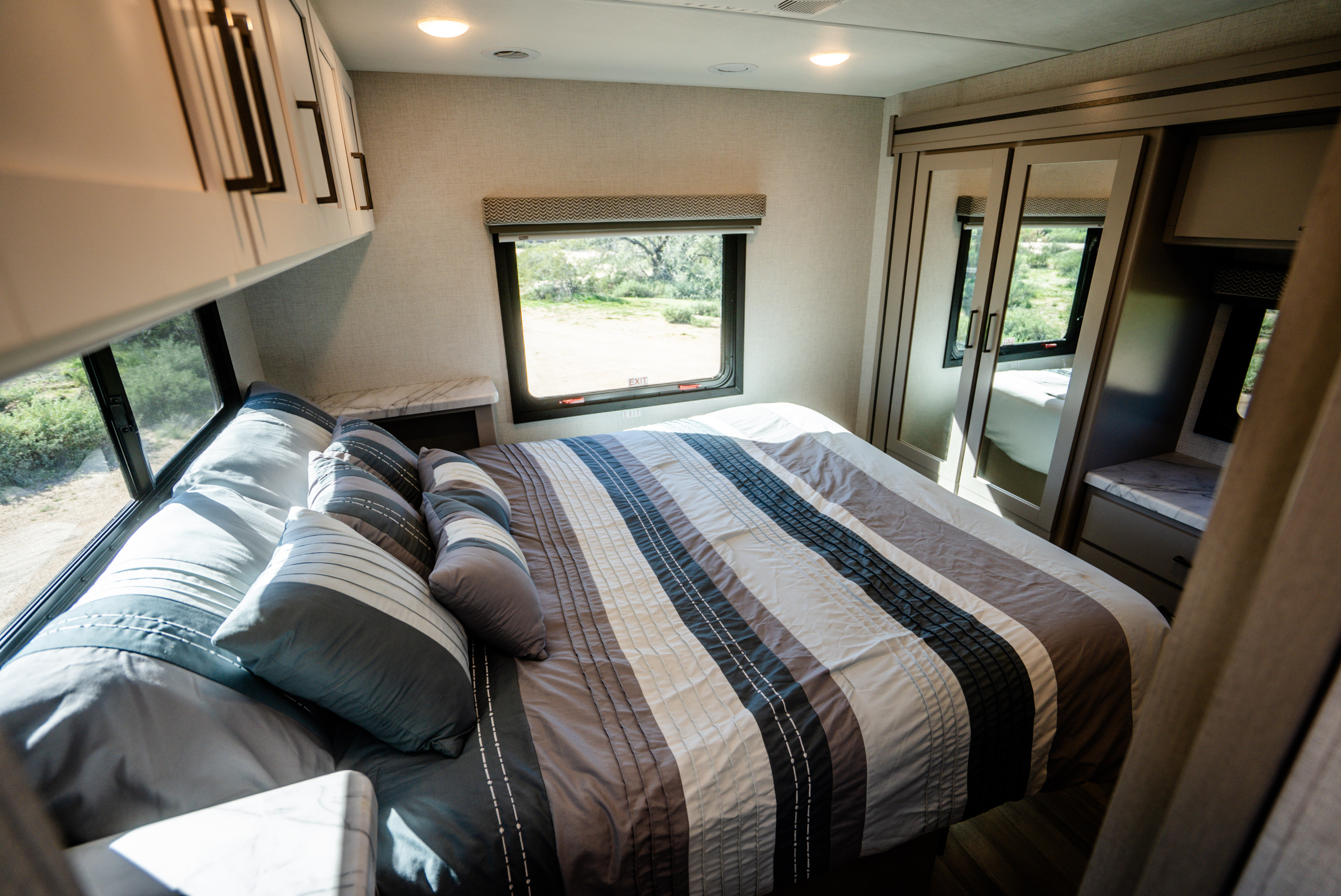 Inside an RV rental bedroom: striped bed, overhead cabinets, and a mirrored closet, with views of nature.. Thor Geneva 2025