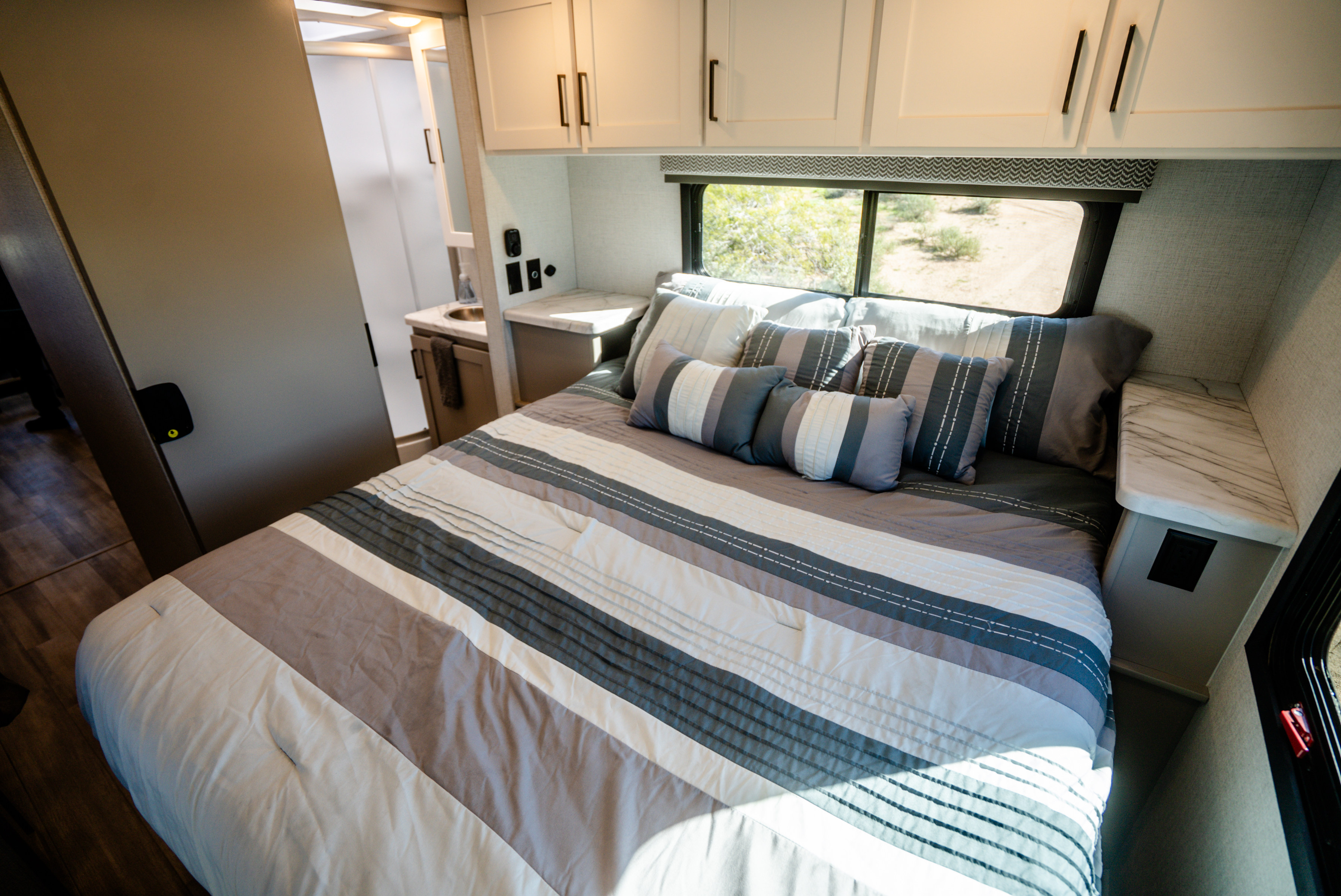 Modern RV rental bedroom with striped bedding, window view, built-in storage, and glimpse of a clean bathroom.. Thor Geneva 2025