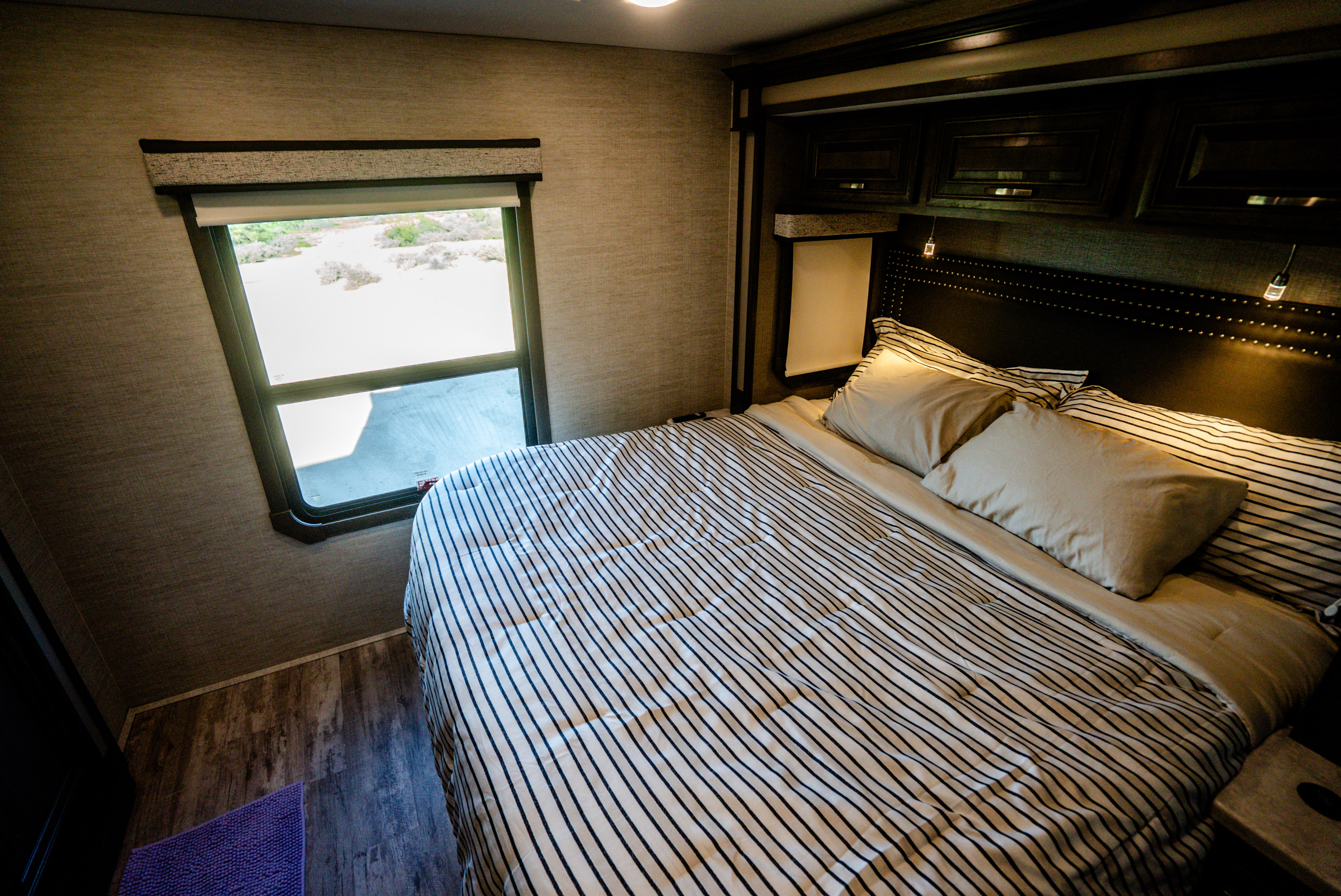 RV rental bedroom showing a bed with striped bedding, light pillows, a window with outdoor view, and dark wood headboard.. Entegra Coach Accolade XL 2023