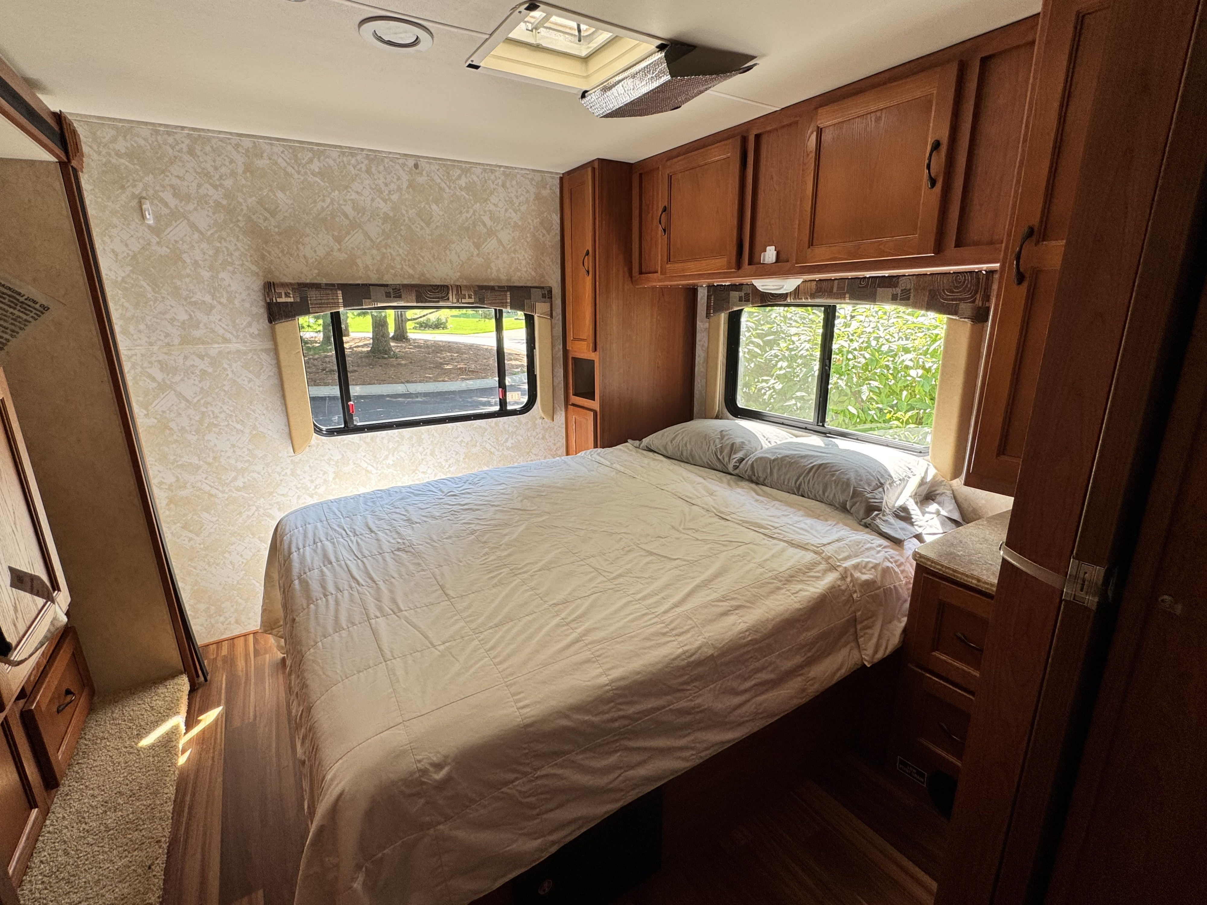 Relaxing RV rental bedroom featuring a large bed, wooden cabinets, overhead storage, and sunlit windows with nature views.. Forest River Coachmen Leprechaun 2013