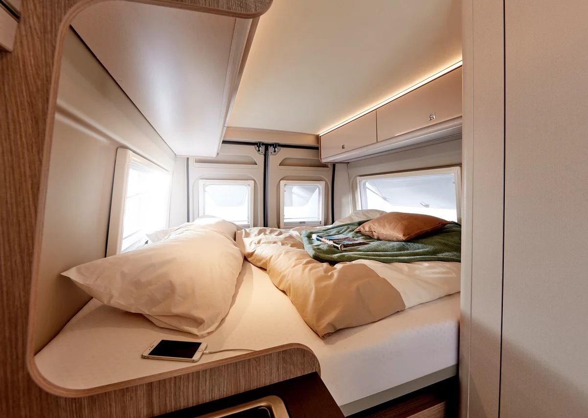 Cozy RV rental bedroom featuring a large bed, bright windows, overhead storage, and a smartphone charging on a bedside shelf.. Other BURSTNER CAMPEO C600 2025