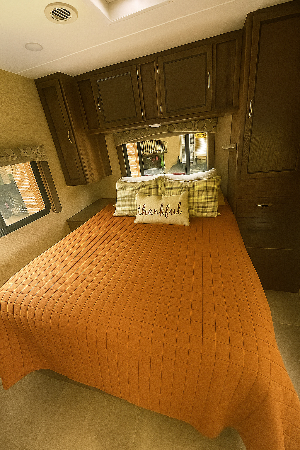 Cozy RV rental bedroom featuring an orange quilted comforter, "thankful" pillow, wood cabinets, and windows.. Coachman Freelander 27QB 2019