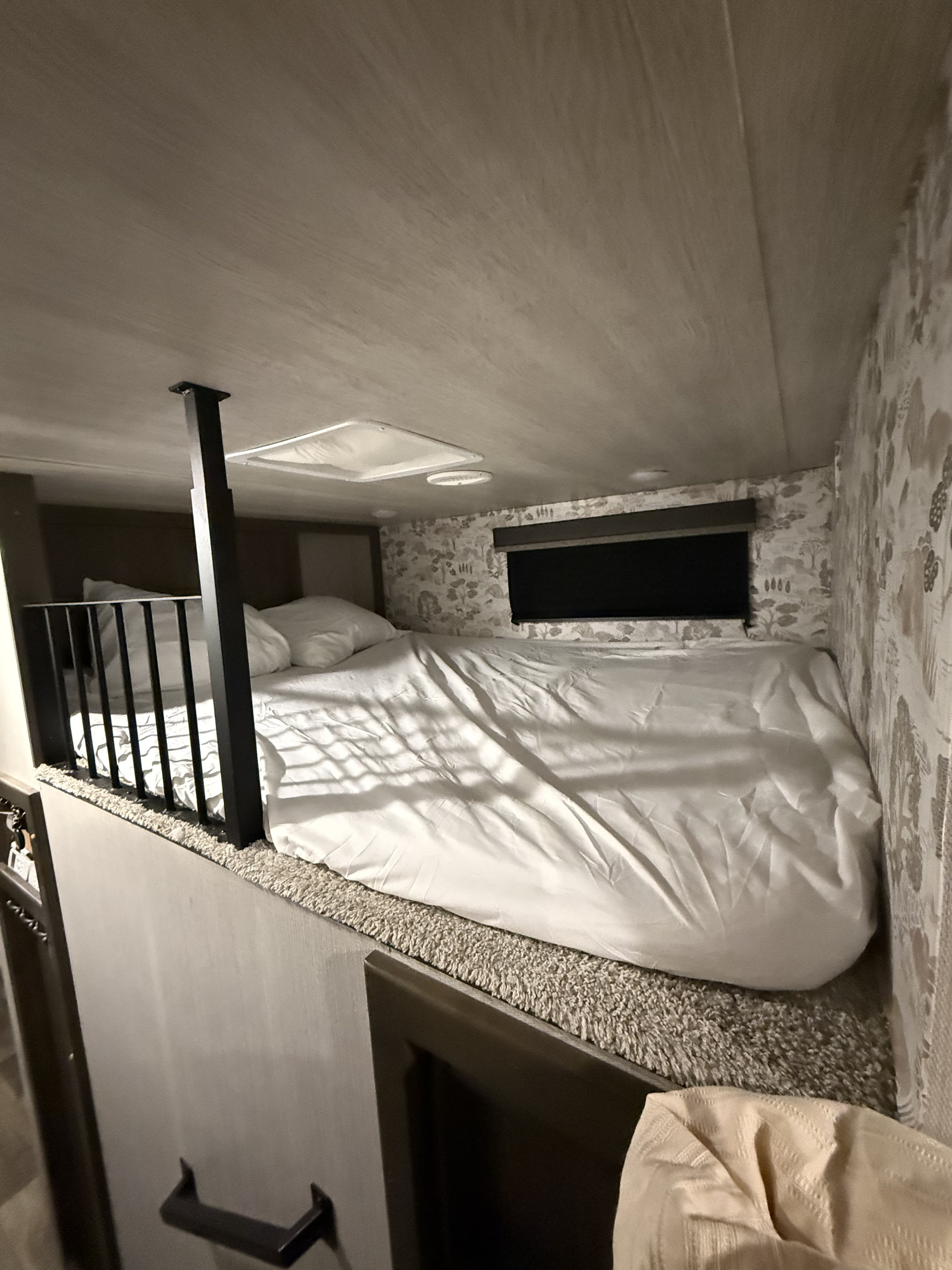 Cozy loft bed with white bedding and a safety railing inside an RV rental. Features patterned wallpaper and a small window.. Keystone RV Cougar 2021