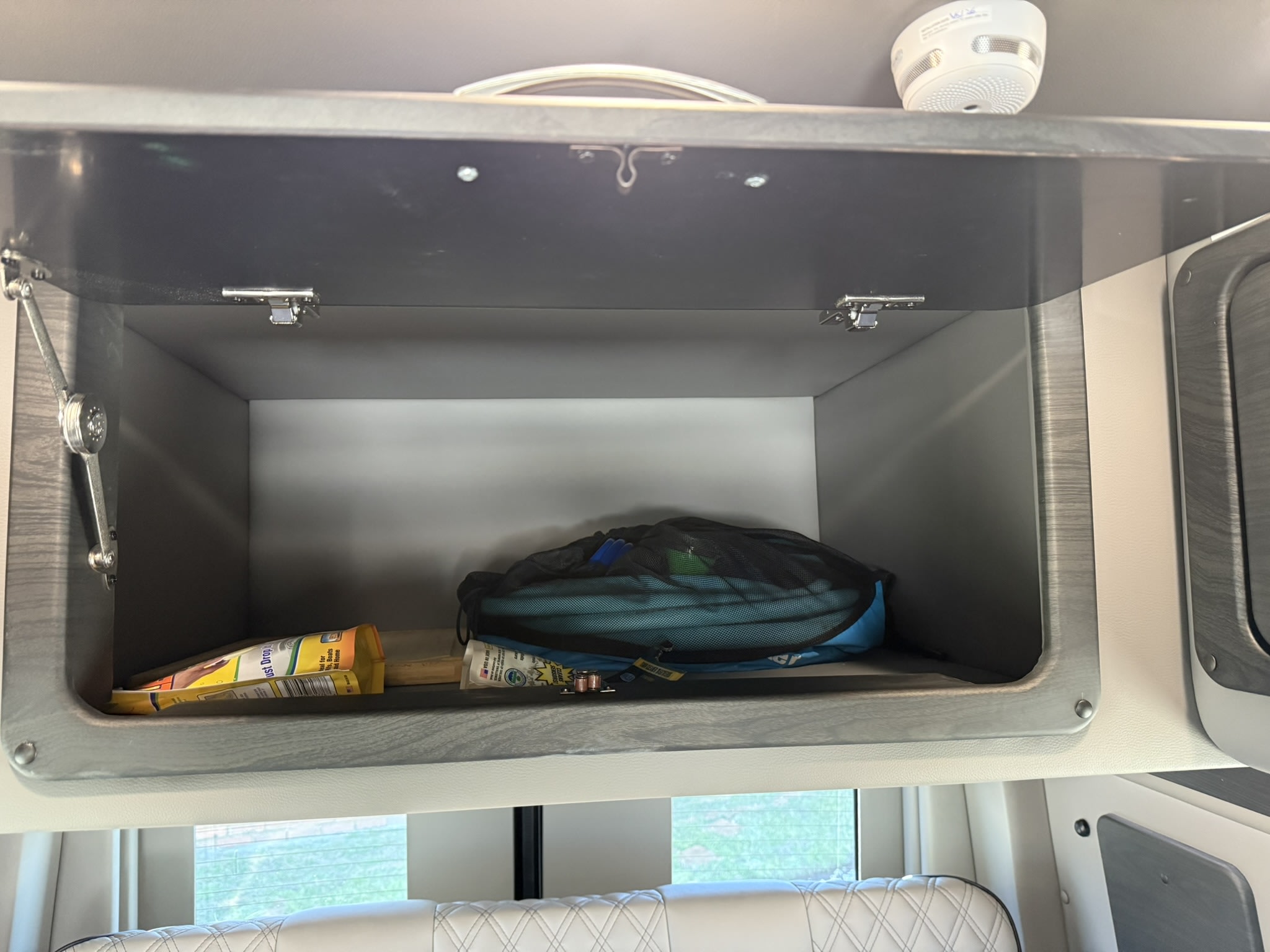 Open overhead storage cabinet in an RV rental. Visible items include a mesh bag, snacks, and small supplies. Great for travel.. Midwest Automotive Designs Ultimate CamperVan 2023