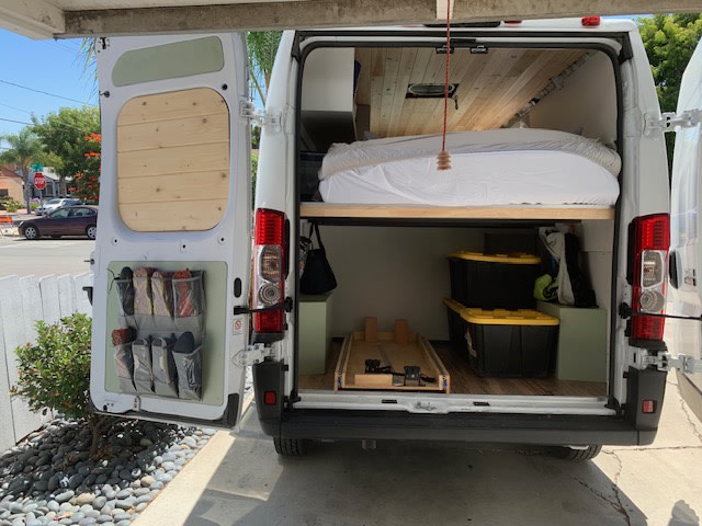 Back of RV rental camper van showing interior: raised bed, storage, and shoe organizer. Custom wood accents visible.. Dodge Ram Promaster 2019