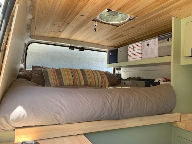 Cozy RV rental's rear bed area. Features a soft bed, wooden plank ceiling, vent, and overhead storage bins.. Dodge Ram Promaster 2019